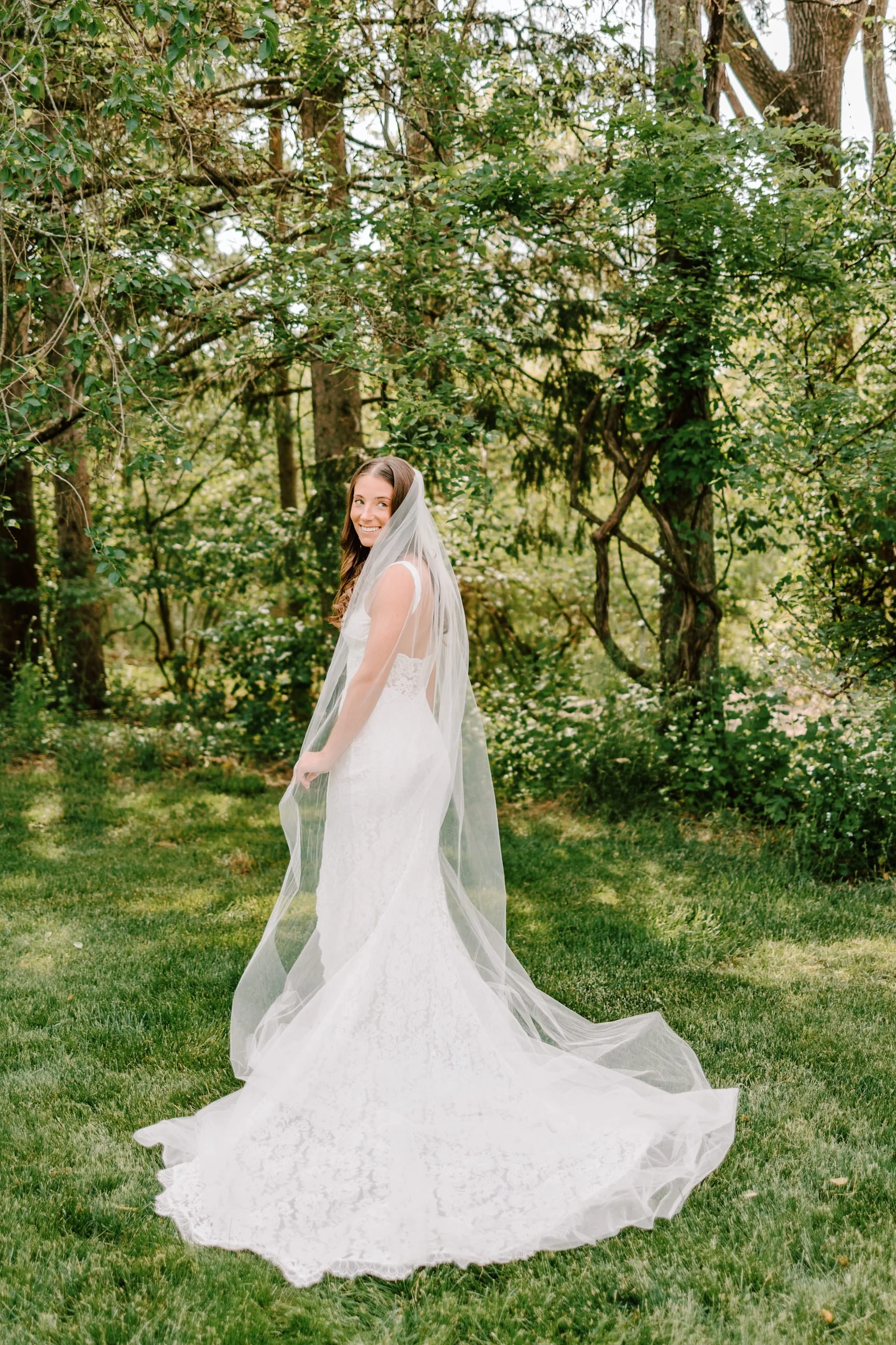 A smiling bride in a white wedding dress and veil standing on grass in a lush, wooded outdoor setting.
