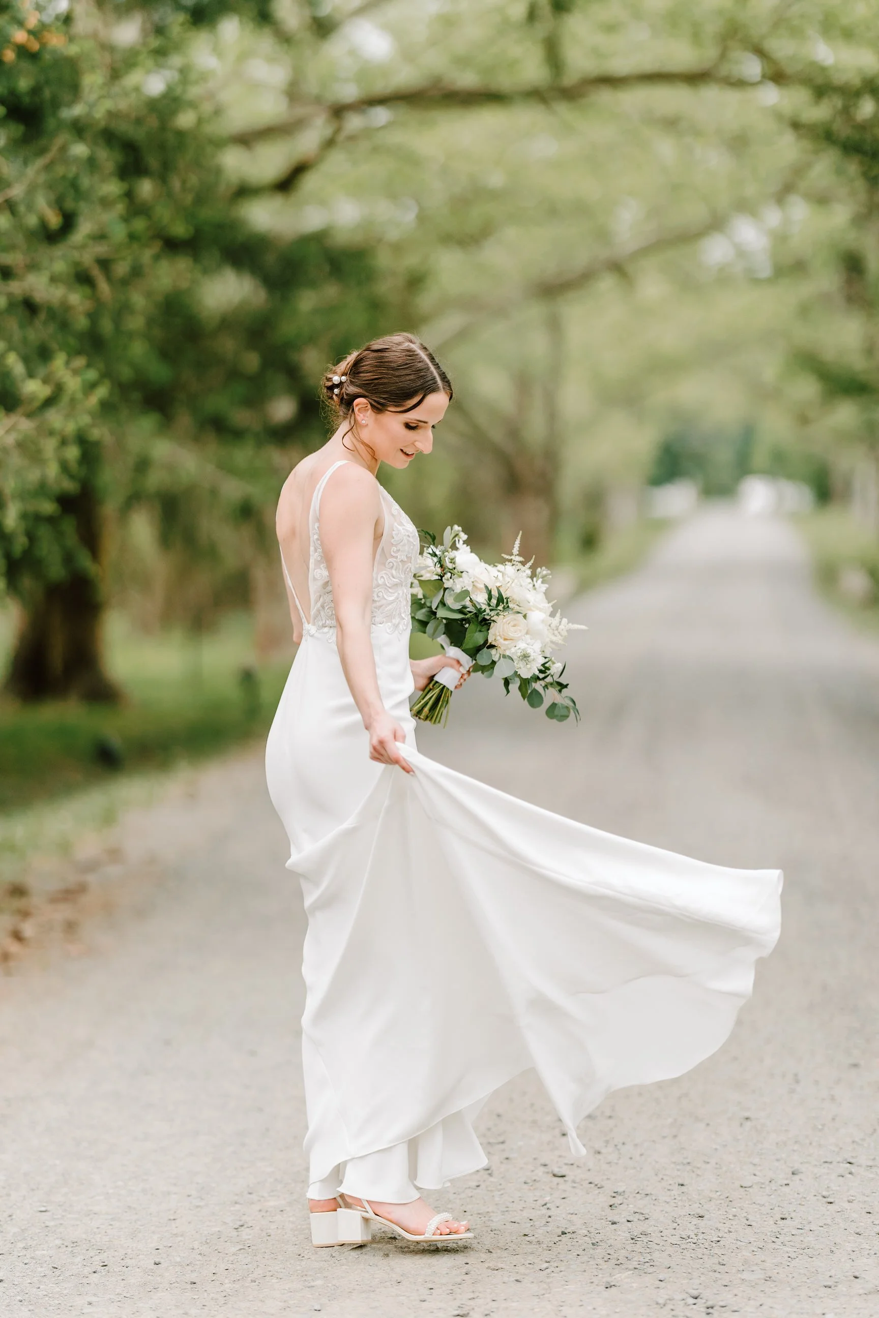 A bride in a white wedding dress holding a bouquet of white flowers, standing on a tree-lined dirt path, lifting her dress slightly and smiling.