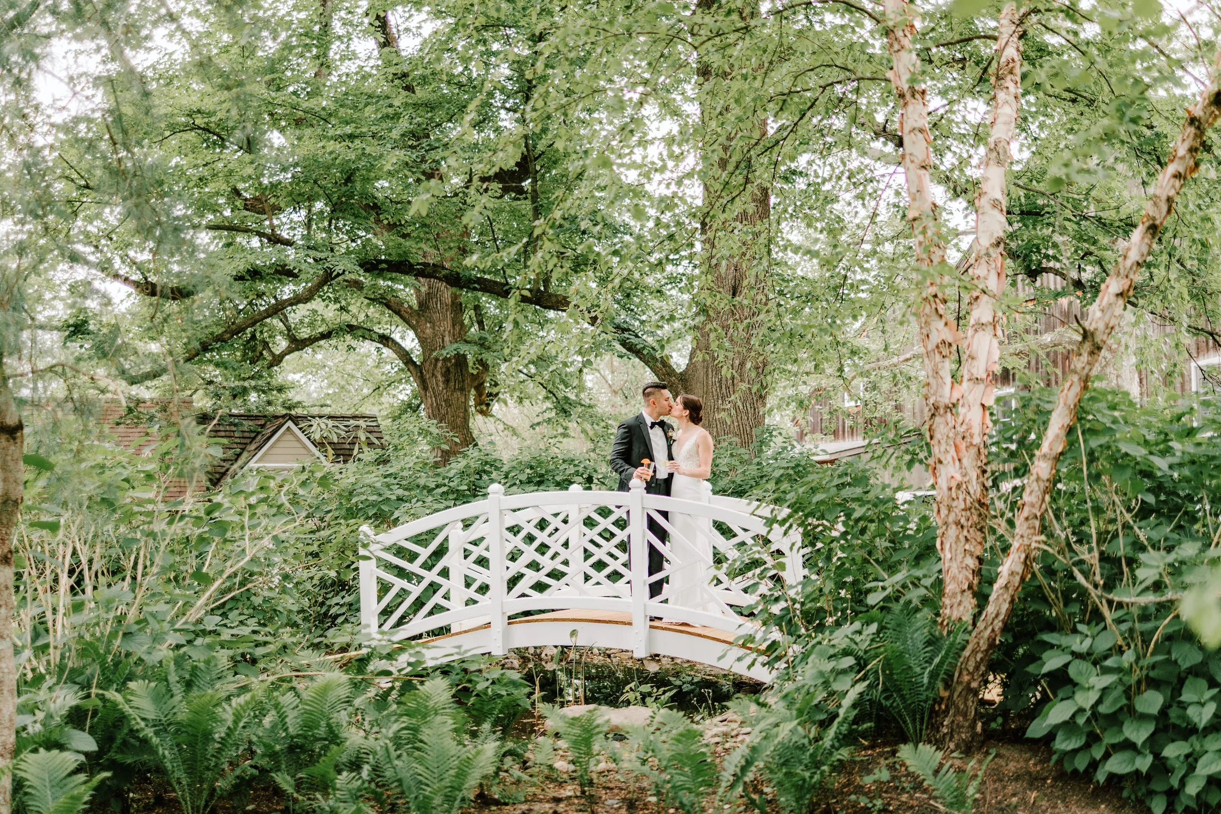 A bride and groom standing on a small white arched bridge in a lush green garden, sharing a kiss.