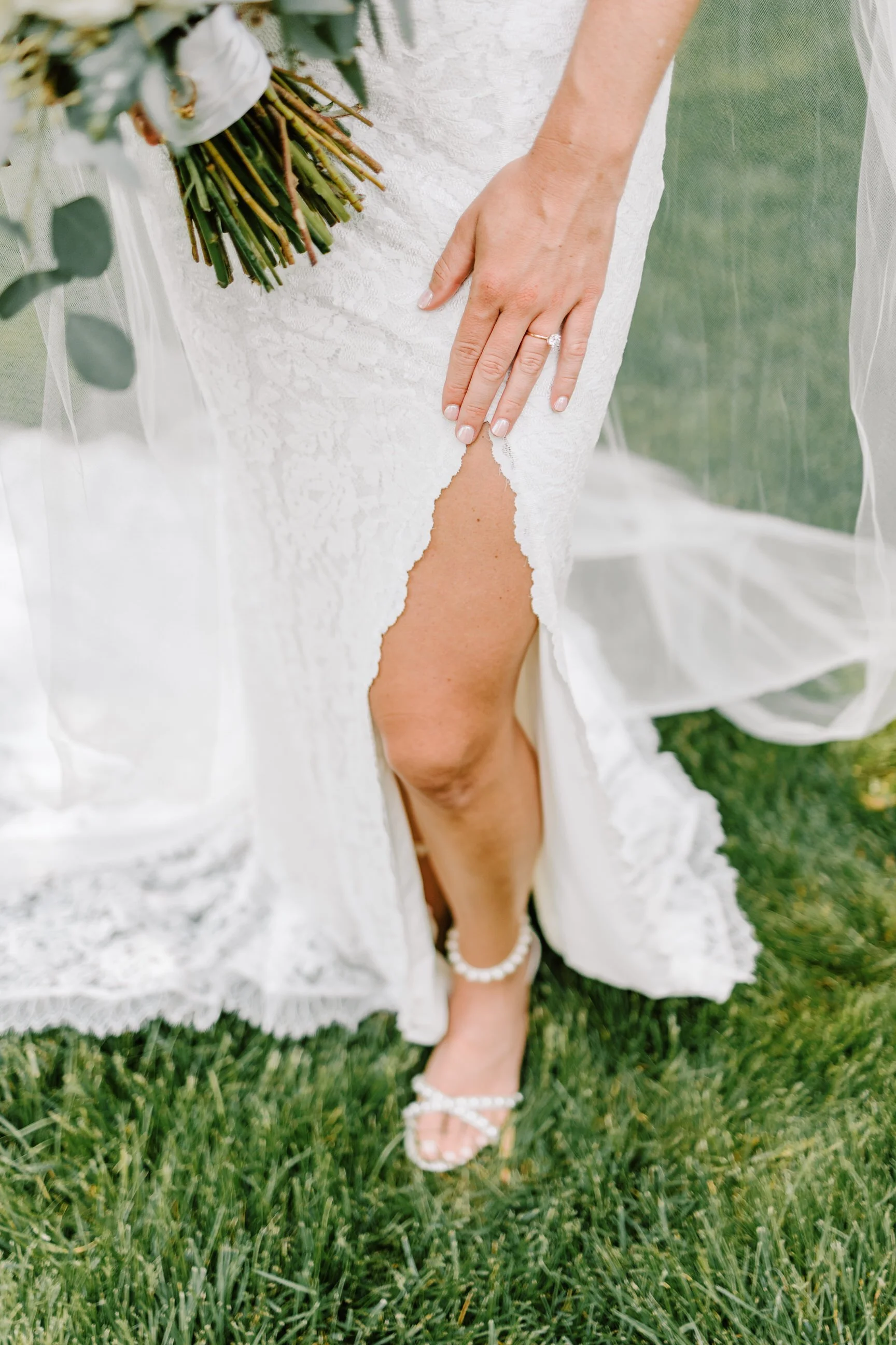 Close-up of a bride's leg showing a slit in her white lace wedding dress, with a hand resting on her thigh showing a wedding ring. She is wearing pearl-embellished heels and holding a bouquet. The dress is slightly lifted to reveal her leg.