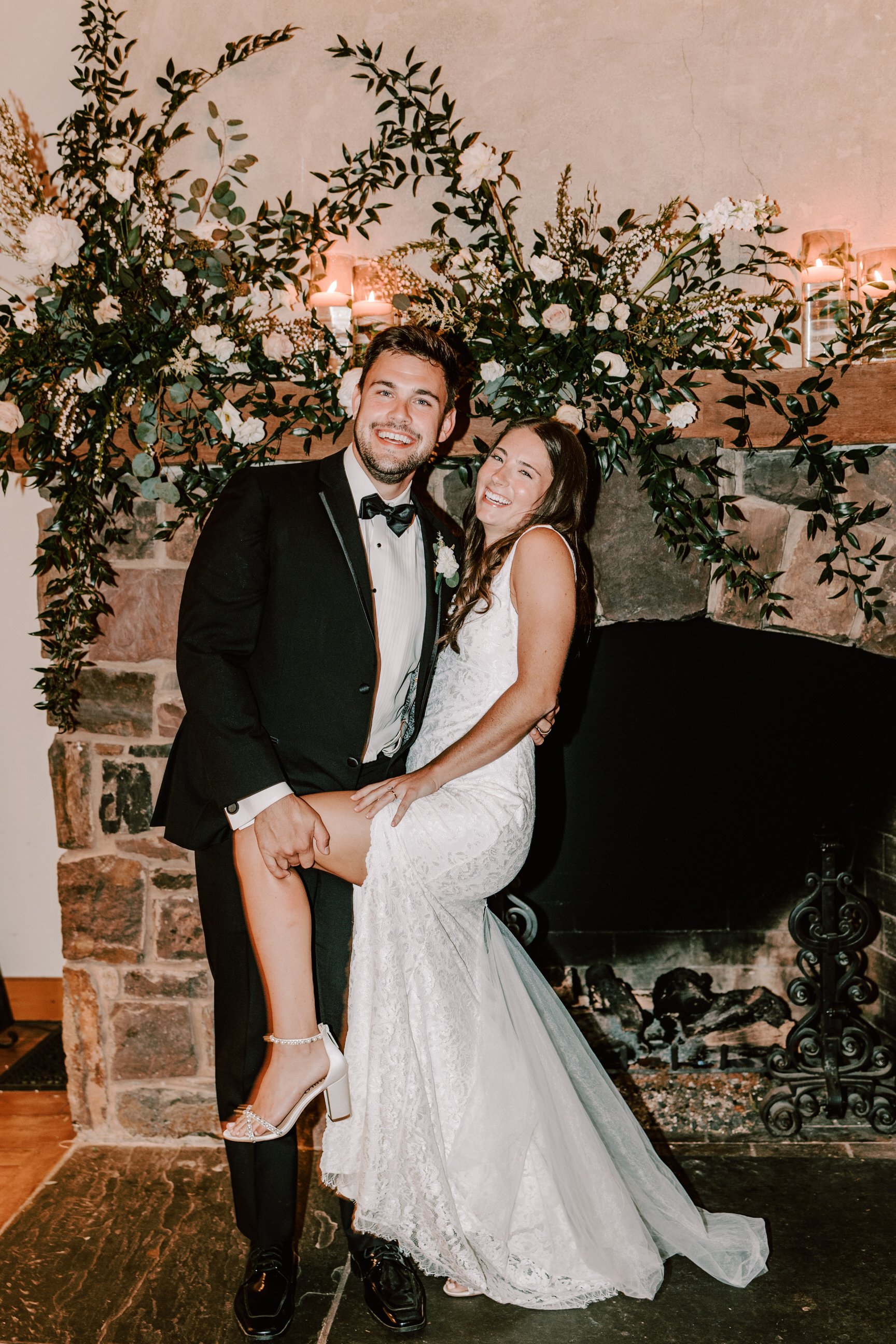 A happy bride and groom celebrating their wedding, with the groom in a tuxedo and the bride in a white wedding dress, standing in front of a fireplace decorated with greenery and flowers.