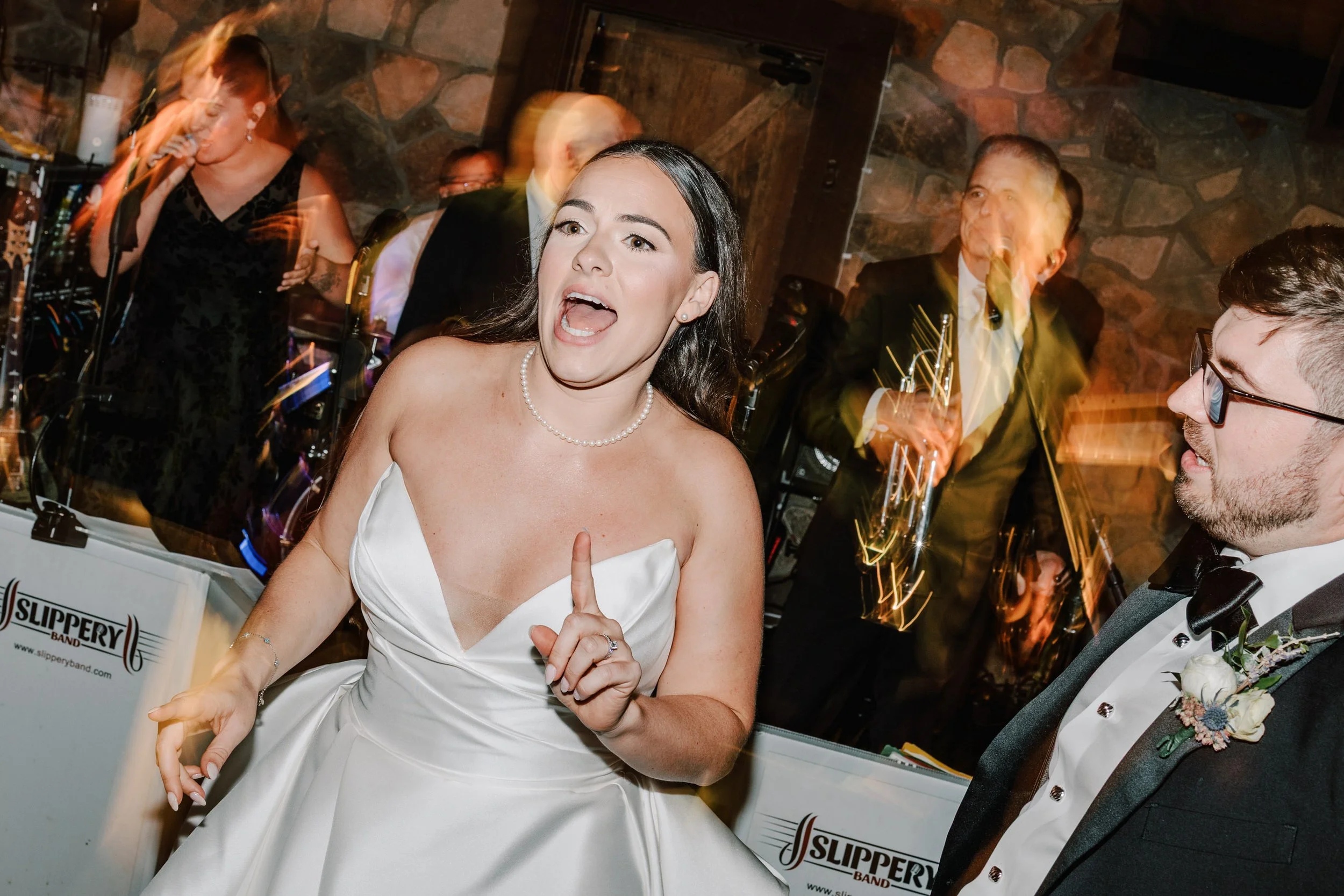 A bride in a white wedding dress singing or speaking confidently at her wedding reception. A man in a tuxedo with glasses stands nearby. In the background, a band is playing with people dancing and enjoying the celebration.
