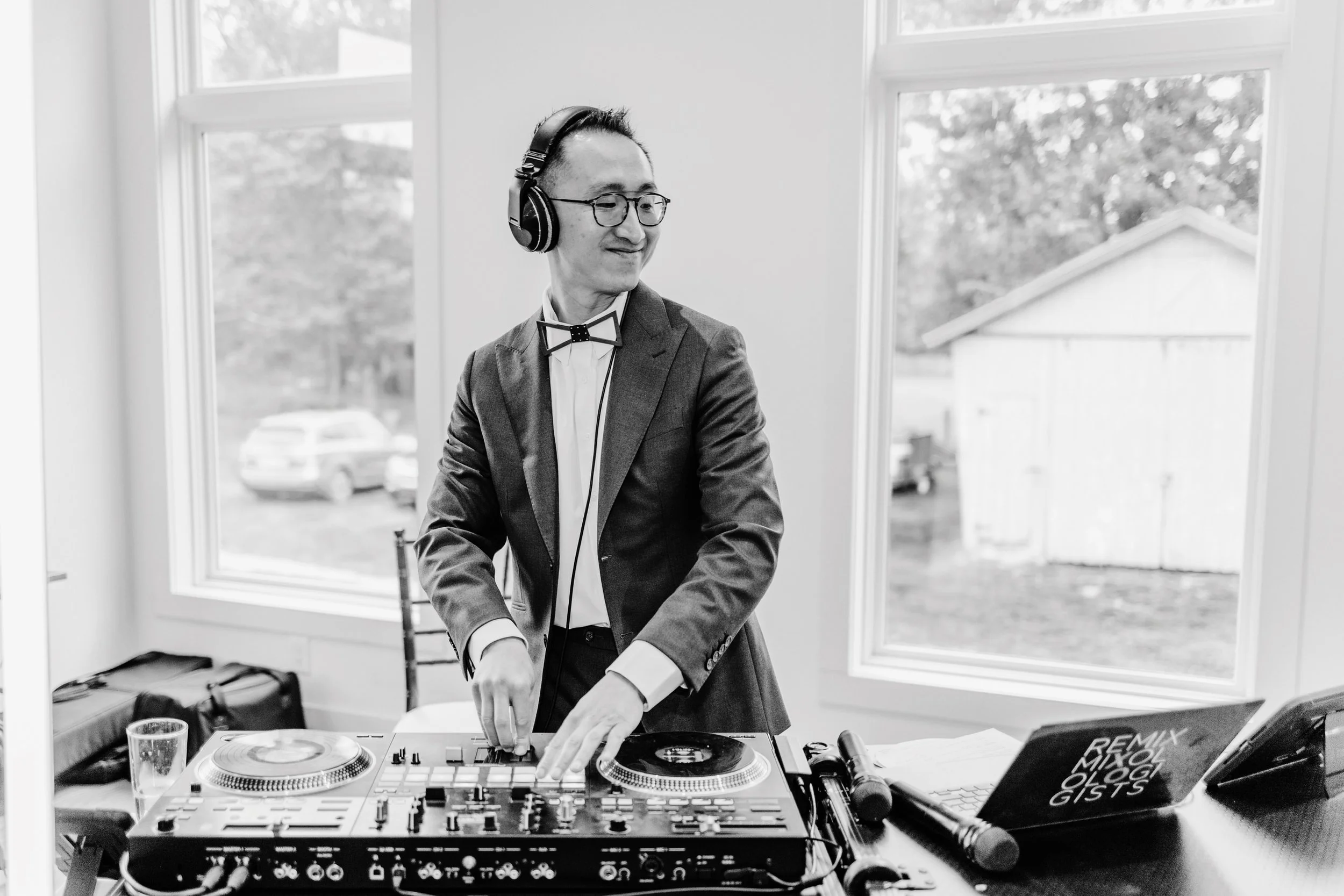 A man in a suit and glasses, wearing a bow tie, DJing with headphones in a bright room with large windows.