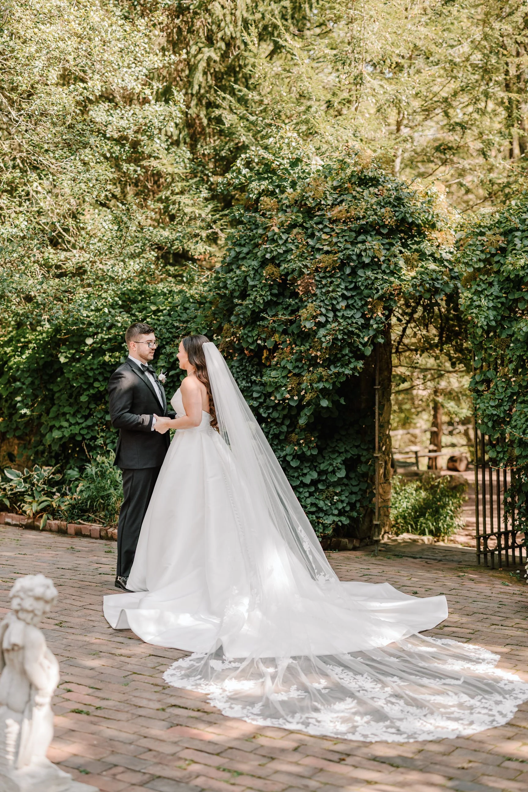 A bride and groom holding hands and facing each other outdoors on a brick path, surrounded by lush green foliage, during their wedding ceremony.