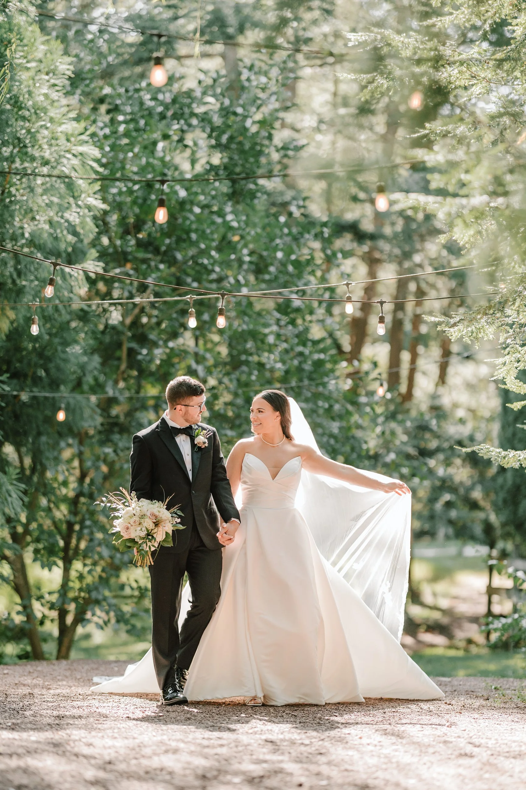 A bride and groom walk hand-in-hand outdoors on their wedding day, surrounded by greenery and string lights, with the bride holding her wedding dress and bouquet, both smiling at each other.