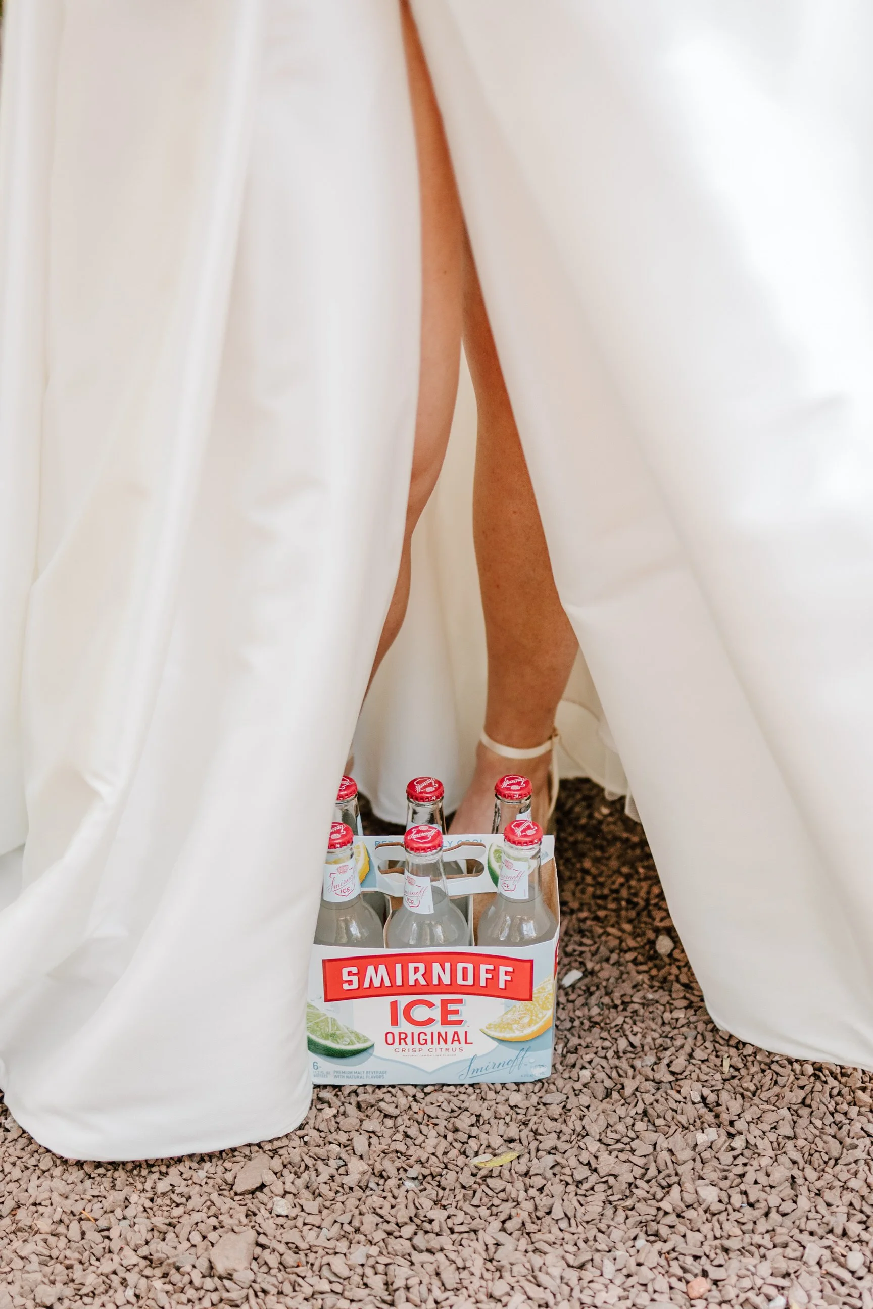 A person standing behind a white curtain with only their legs visible, wearing a white strappy sandal. On the ground, there is a six-pack of Smirnoff Ice Original bottles with red caps and labels, sitting on a gravel surface.