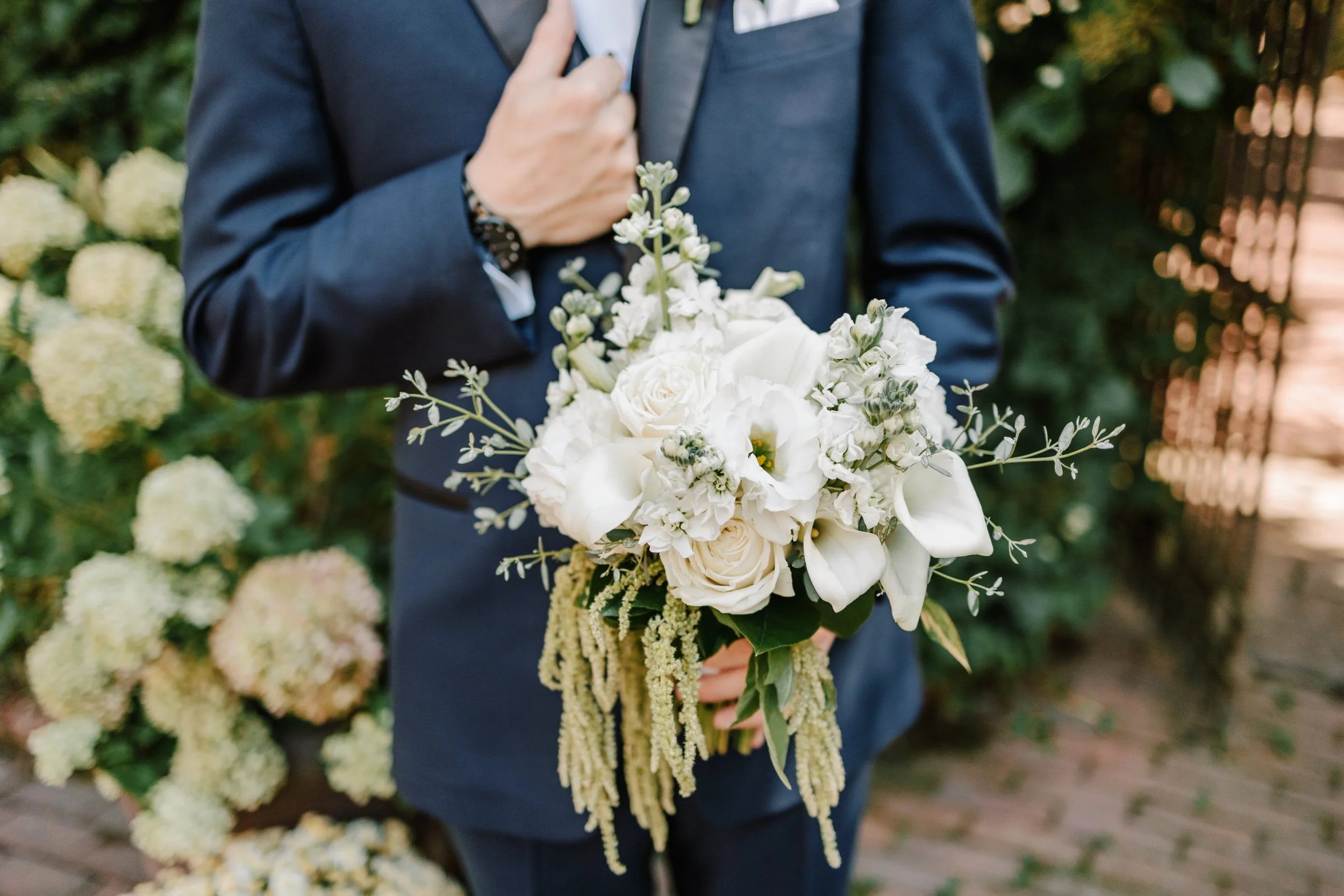 A man in a navy suit holding a bouquet of white roses, calla lilies, and other white flowers, with greenery, outdoors near a brick pathway and floral background.