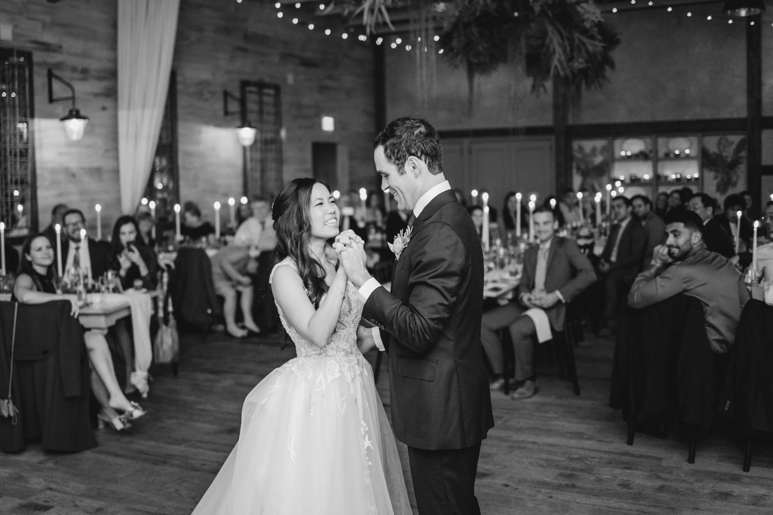 Black and white photo of a bride and groom dancing at their wedding reception, surrounded by seated guests in a decorated venue.