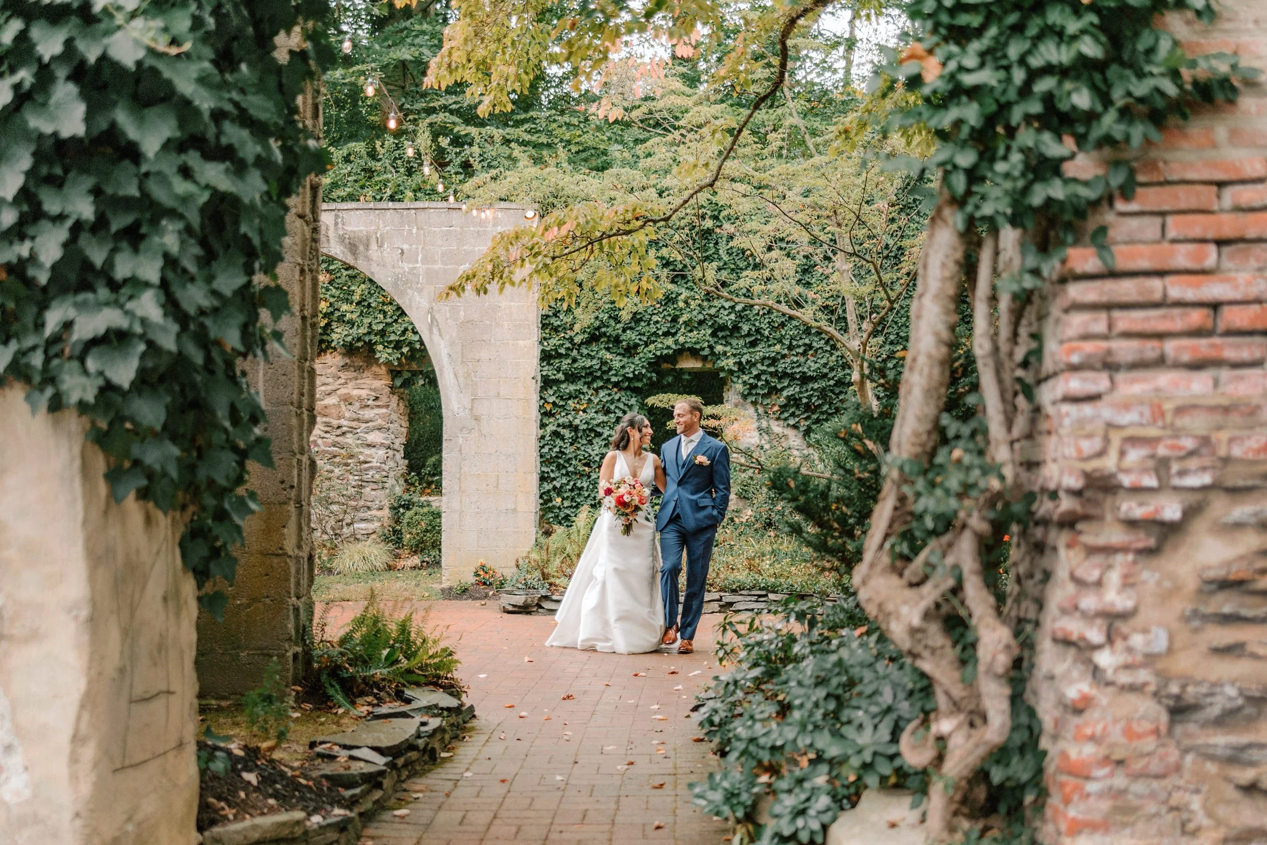 A bride and groom walking through a garden with ivy-covered walls and archways. The bride is wearing a white gown and holding a bouquet, while the groom is in a blue suit. The path is lined with greenery and trees.