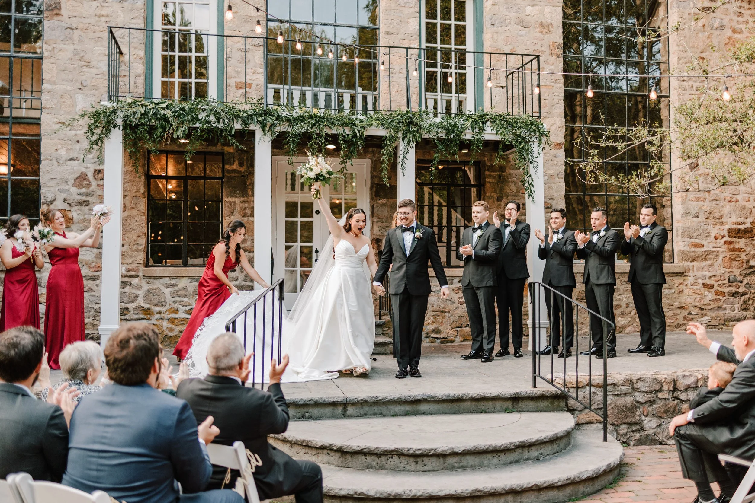 A wedding celebration outside a rustic stone building with string lights overhead. The bride in a white gown is holding a bouquet and raising her arm in celebration, holding hands with the groom in a black tuxedo. Bridesmaids in red dresses and groom