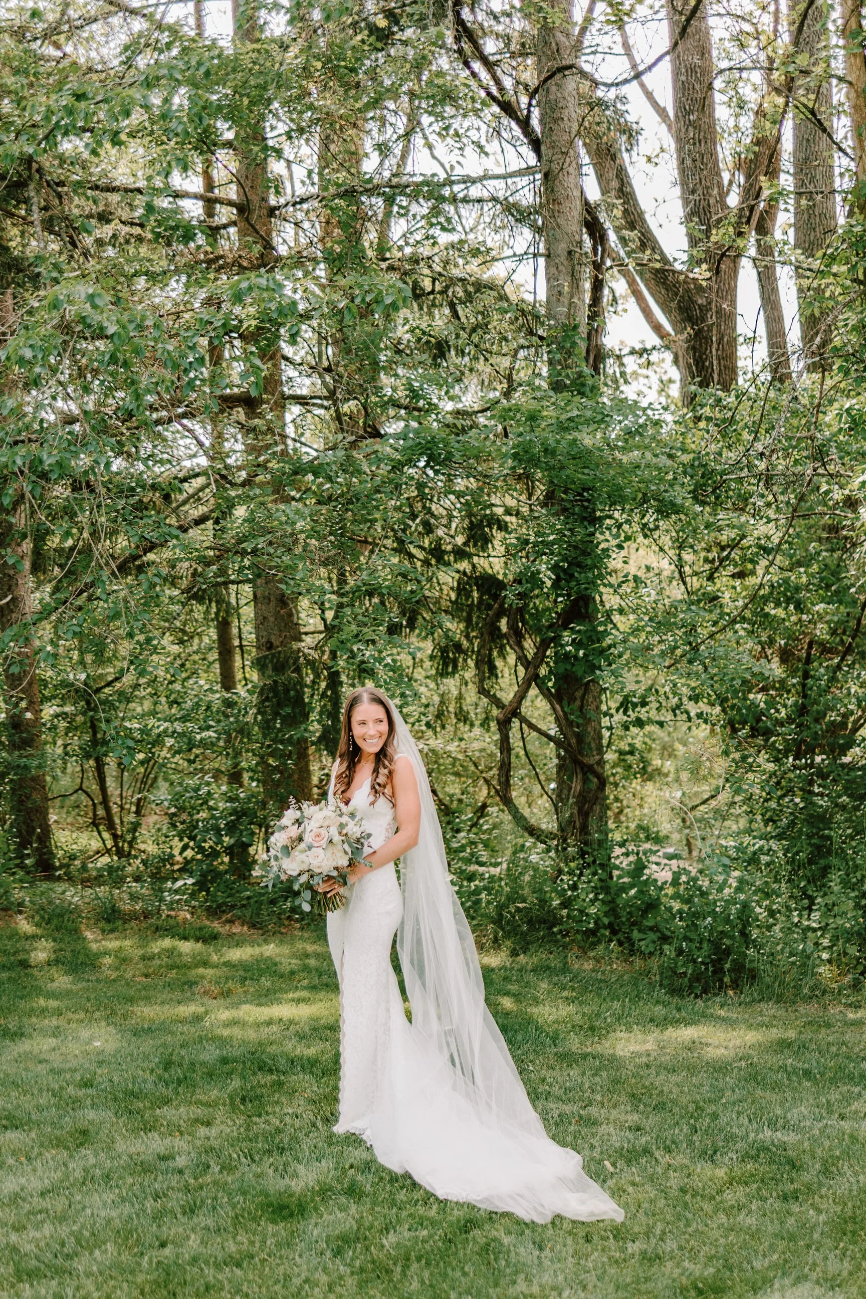 A smiling bride in a white wedding dress holding a bouquet of flowers, standing on grass in a wooded outdoor setting with trees.