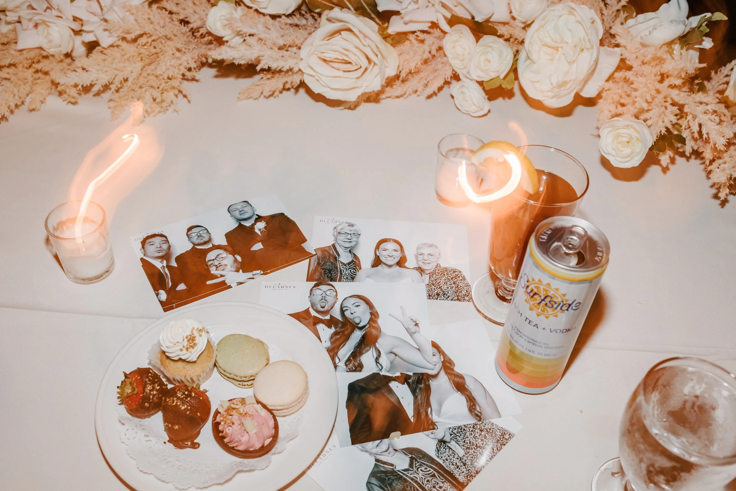 A table decorated with a floral arrangement, photos of people, glasses of drinks, a can of sparkling ice beverage, and a plate with cookies and strawberries.