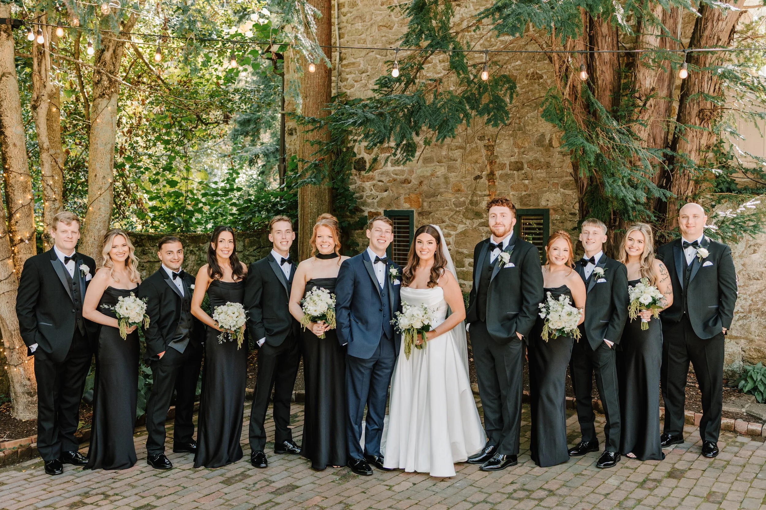 A wedding party standing outdoors in front of a stone wall with trees and string lights. The group includes the bride and groom in the center, surrounded by bridesmaids and groomsmen dressed in formal black and navy attire, holding bouquets and weari