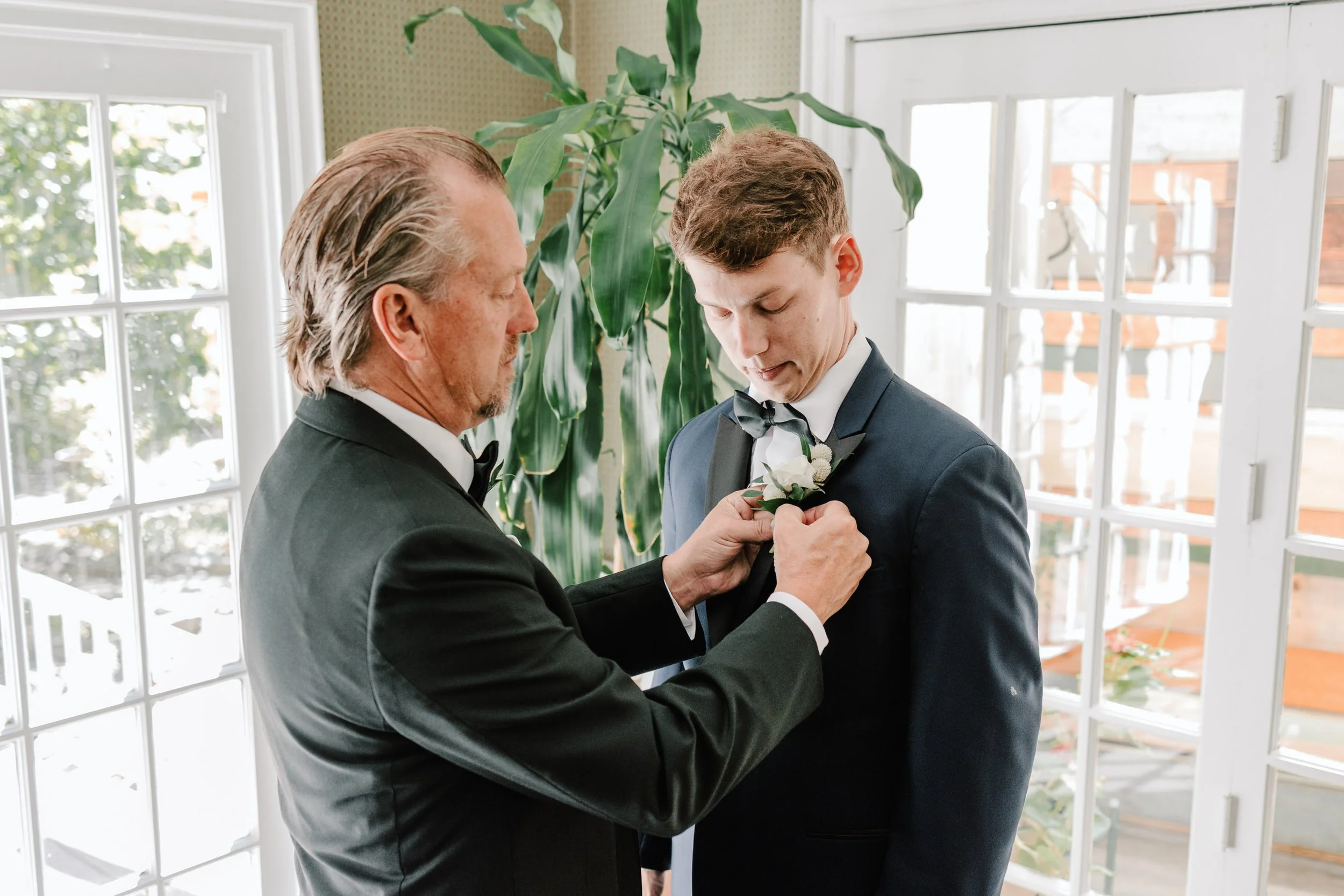 An older man helping a young man with a boutonniere for a wedding, both dressed in tuxedos, in a bright room with large windows and green plants.