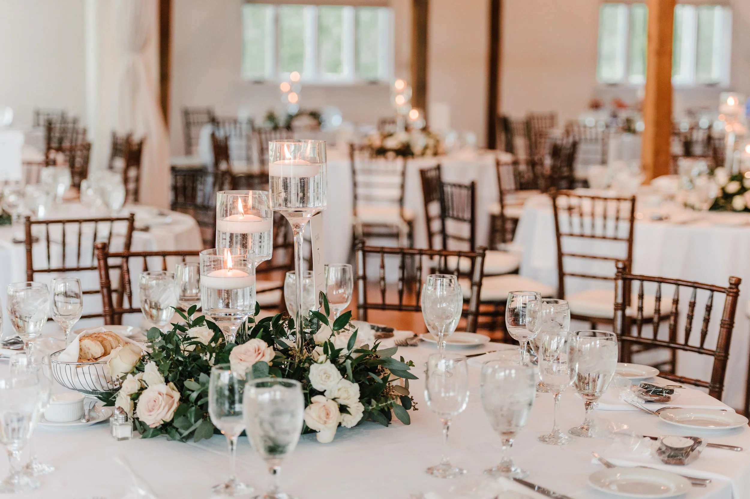 Wedding reception table with white tablecloth, floral centerpiece, and tall glass candle holders with floating candles, set with glassware, plates, silverware, and bread, in a decorated hall with wooden beams and windows.