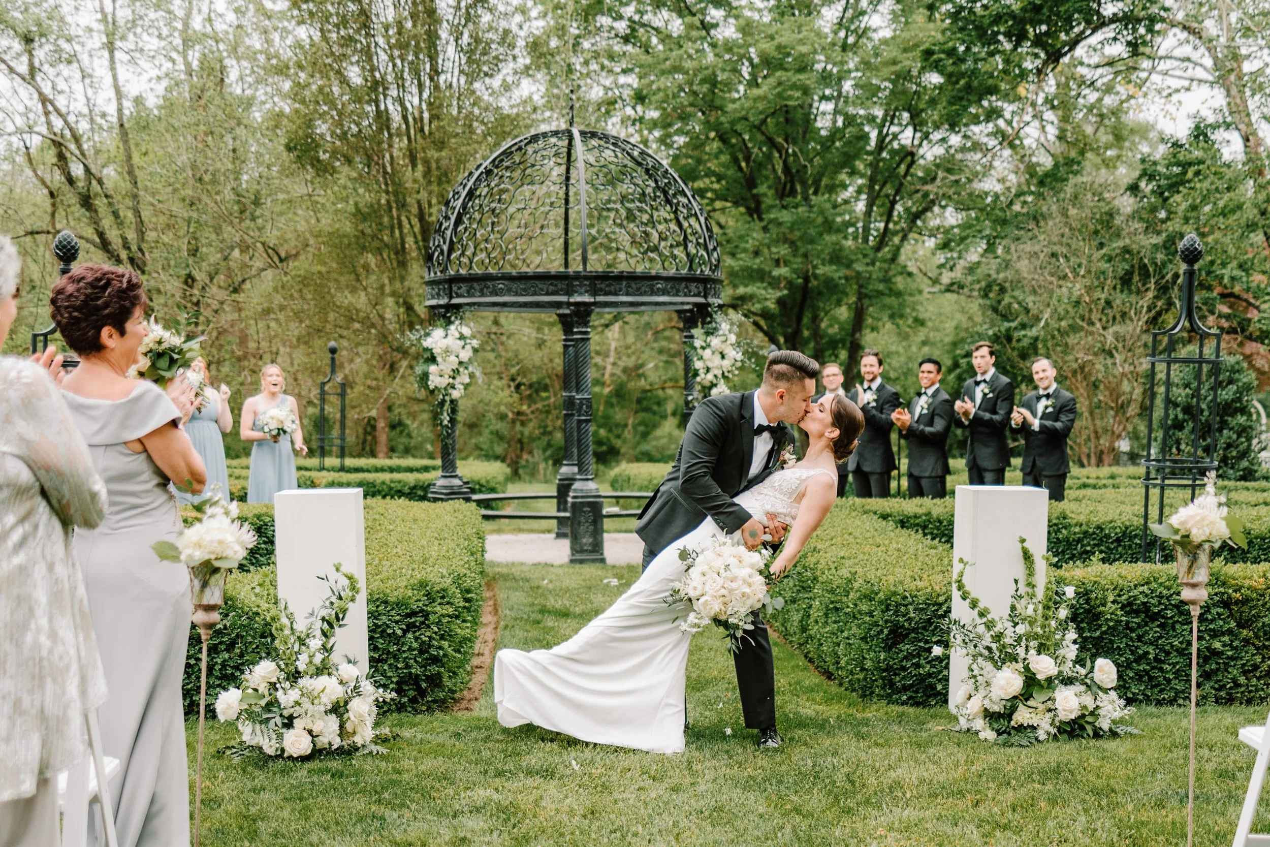 A wedding ceremony outdoors with a bride and groom sharing a kiss, surrounded by friends and family, with a green garden and decorative arch in the background.