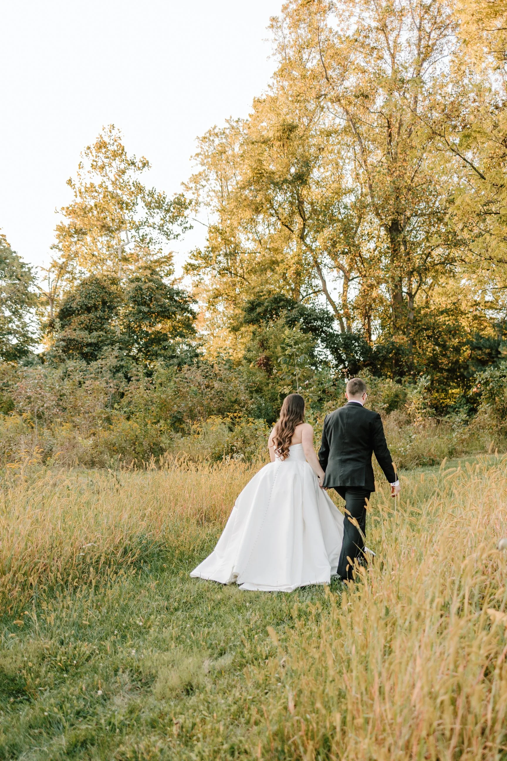 A bride and groom walk hand in hand through a grassy field with trees in fall foliage in the background.