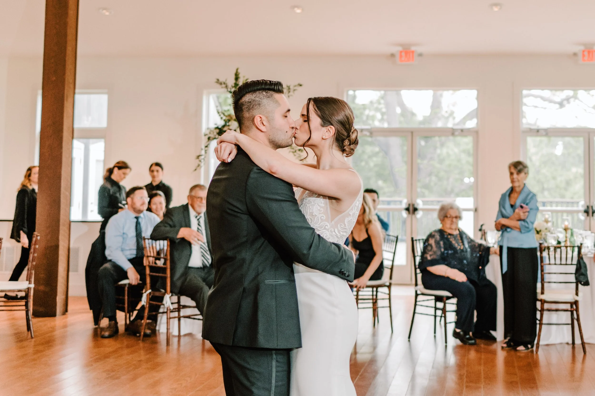 A bride and groom kiss during their wedding dance in a decorated reception hall with guests watching.