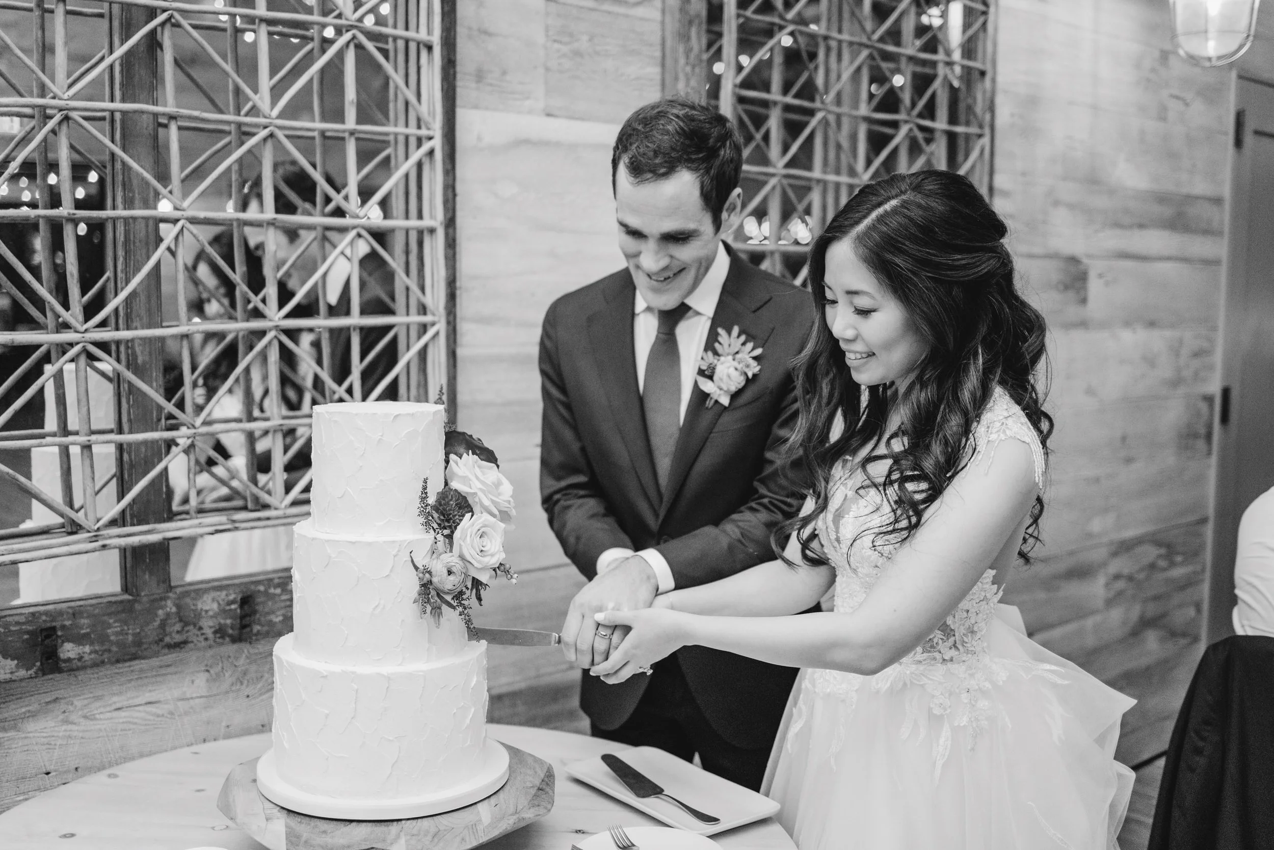A bride and groom cutting a wedding cake together, smiling, in a rustic setting with wooden and metal decor.