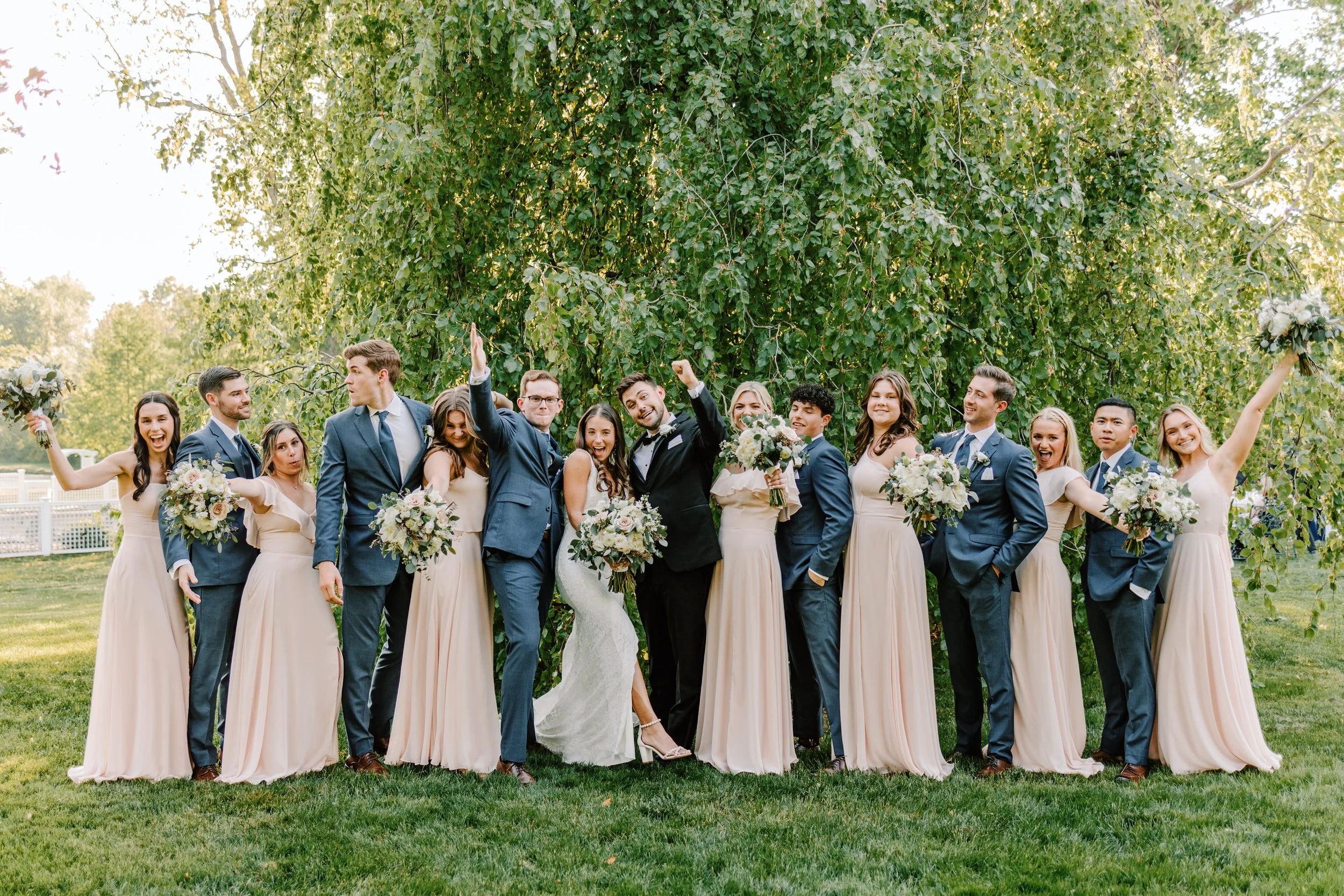A group of people, including a bride and groom, celebrating outdoors in front of a large tree, dressed in wedding attire with the women in light pink dresses and the men in suits, holding bouquets of flowers, and smiling at the camera.