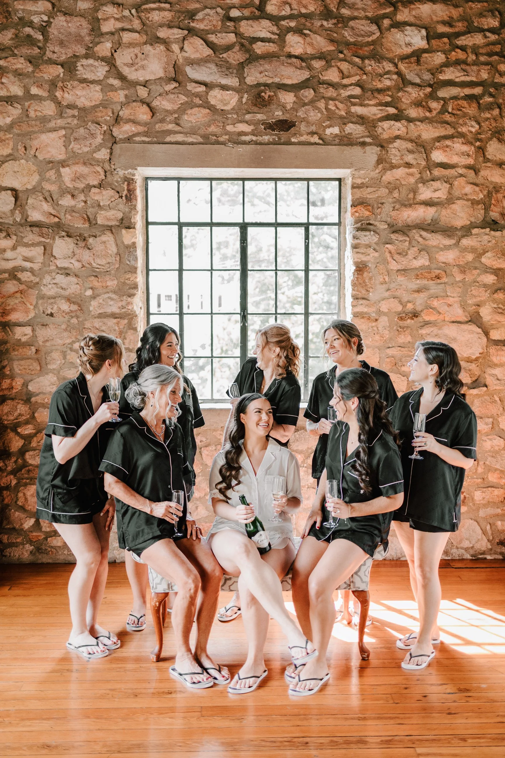 A group of women in matching black pajamas with white piping, celebrating with glasses of champagne and a bottle, in front of a large window with sunlight streaming in, inside a rustic brick-walled room with wooden floors.