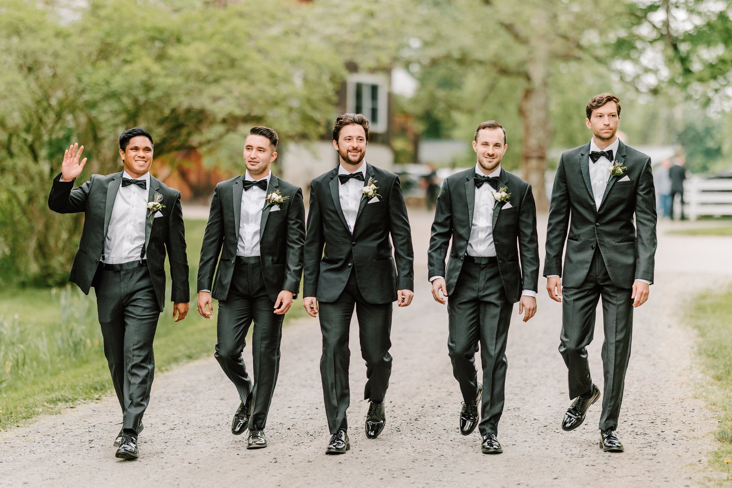 A group of five men dressed in black tuxedos with white shirts and black bow ties, walking outdoors on a gravel path surrounded by greenery, with one man waving and smiling.