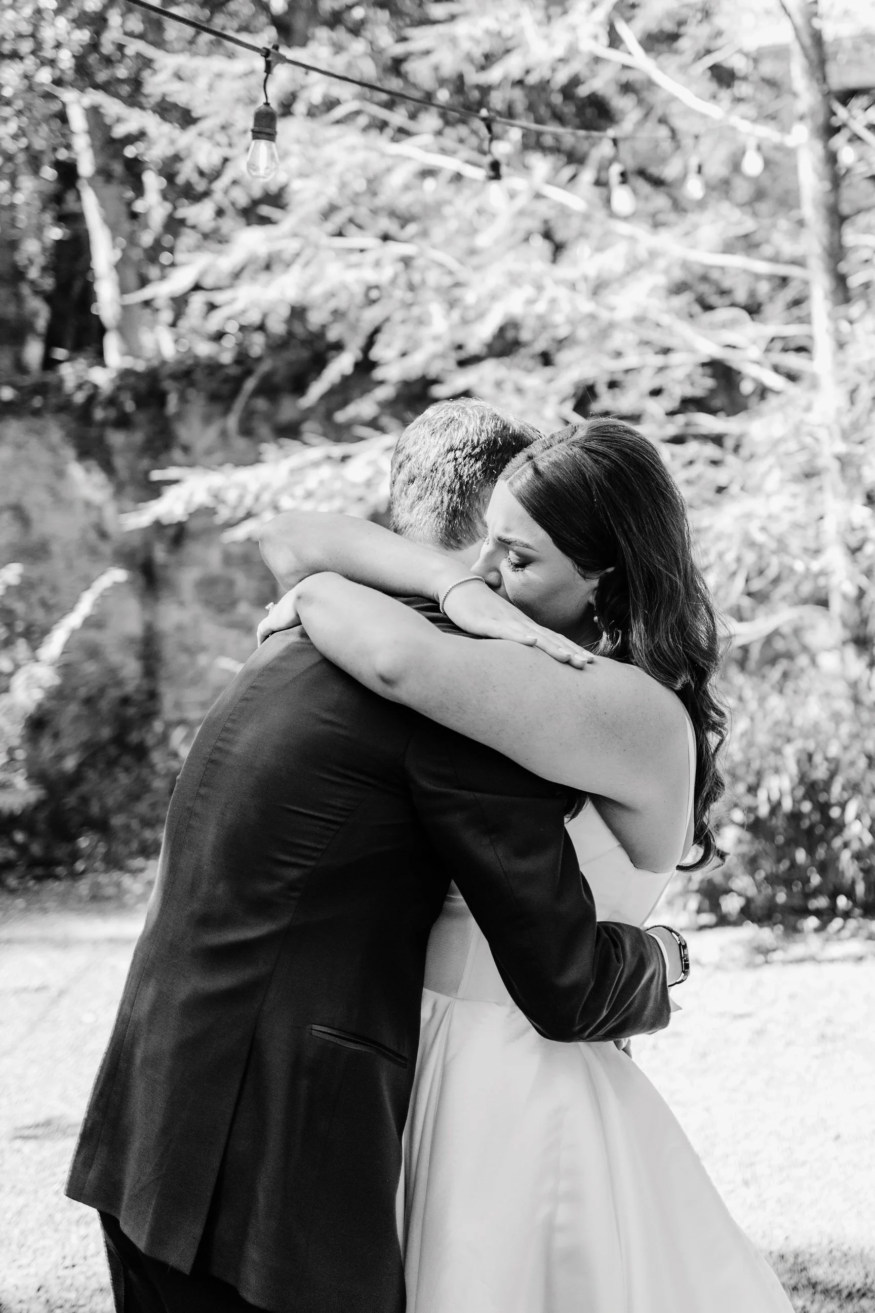 A black and white photograph of a couple embracing tightly. The woman has her eyes closed and appears emotional. They are outdoors with trees in the background and string lights hanging above them.