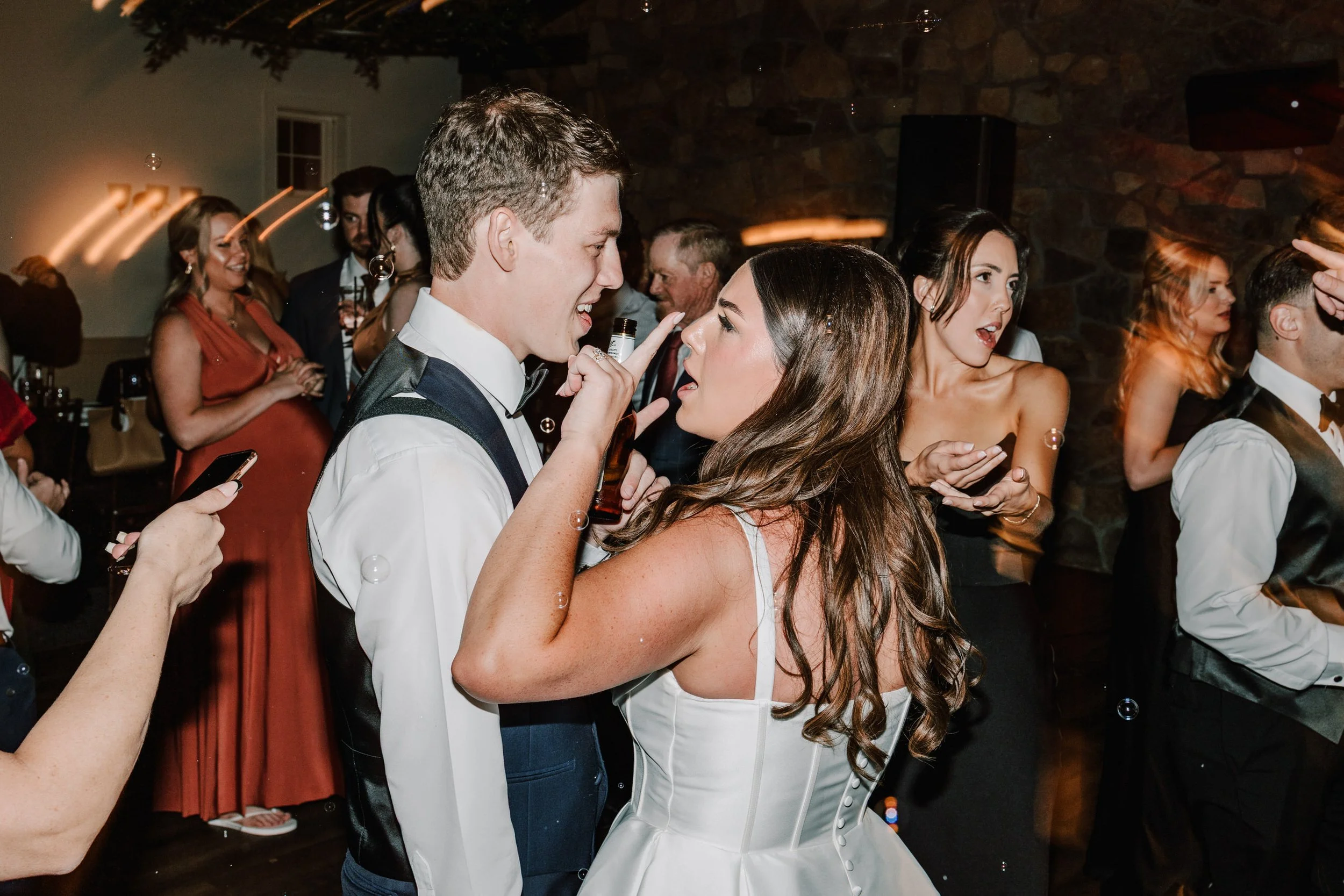 Couple dancing and having fun at wedding reception, brunette woman in white dress and young man in tuxedo, surrounded by other guests.