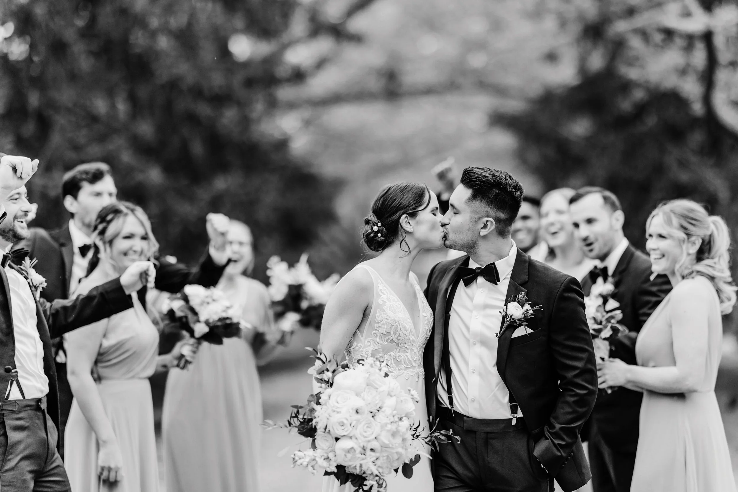 Black and white photo of a newlywed couple kissing during their wedding ceremony, surrounded by smiling friends and family holding bouquets, outdoors with trees in the background.