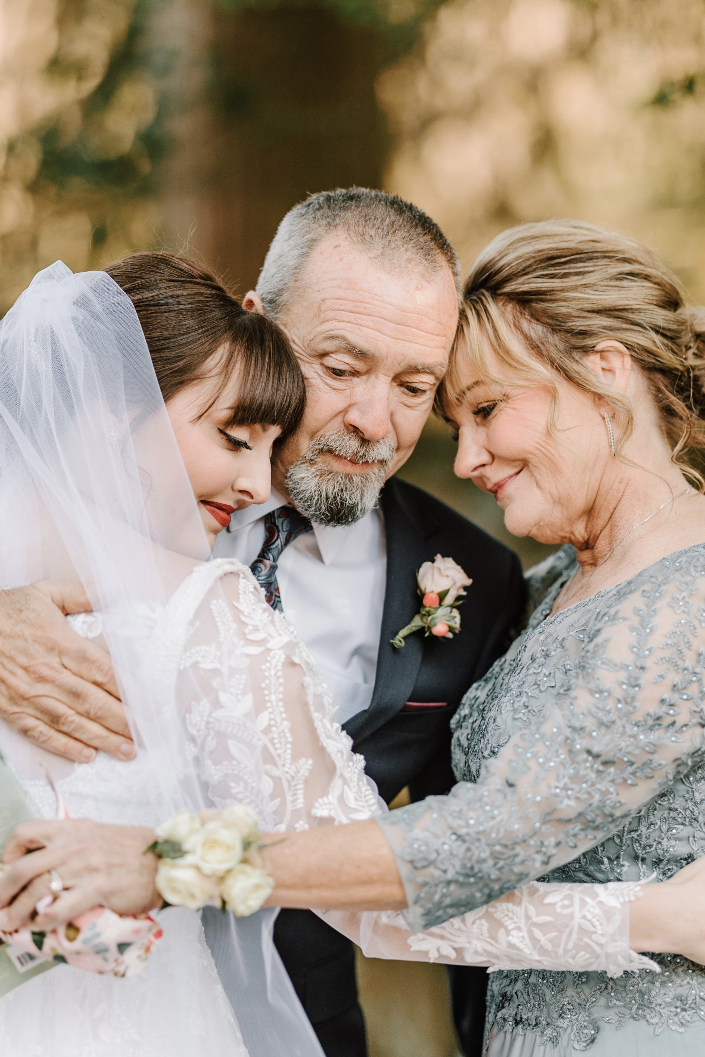 A bride in a white lace dress and veil embraces an older man in a suit and an older woman in a blue embellished gown, all smiling affectionately.