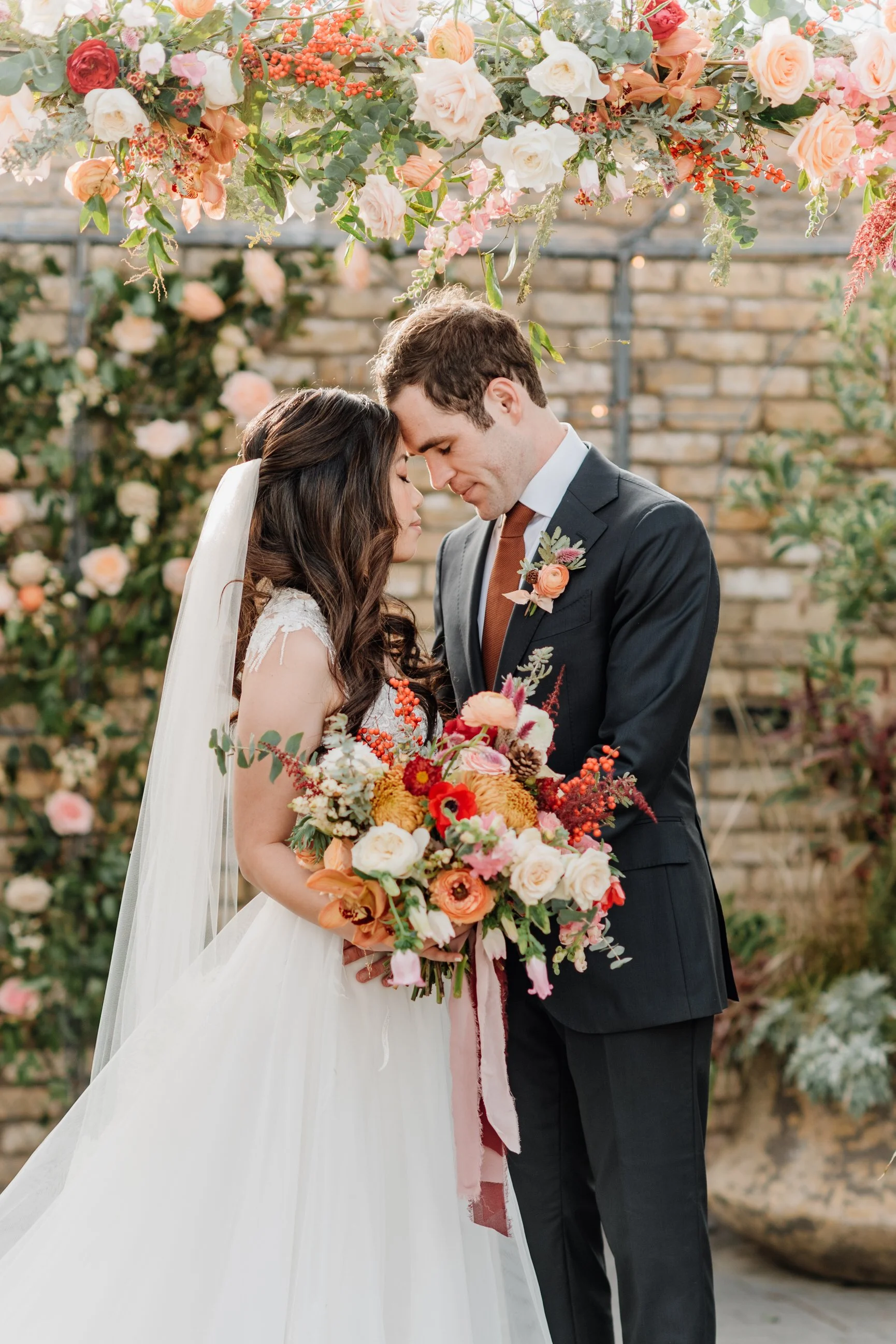 A bride and groom share a tender moment with their foreheads touching during their wedding ceremony, surrounded by a floral arch and arrangements of colorful flowers.
