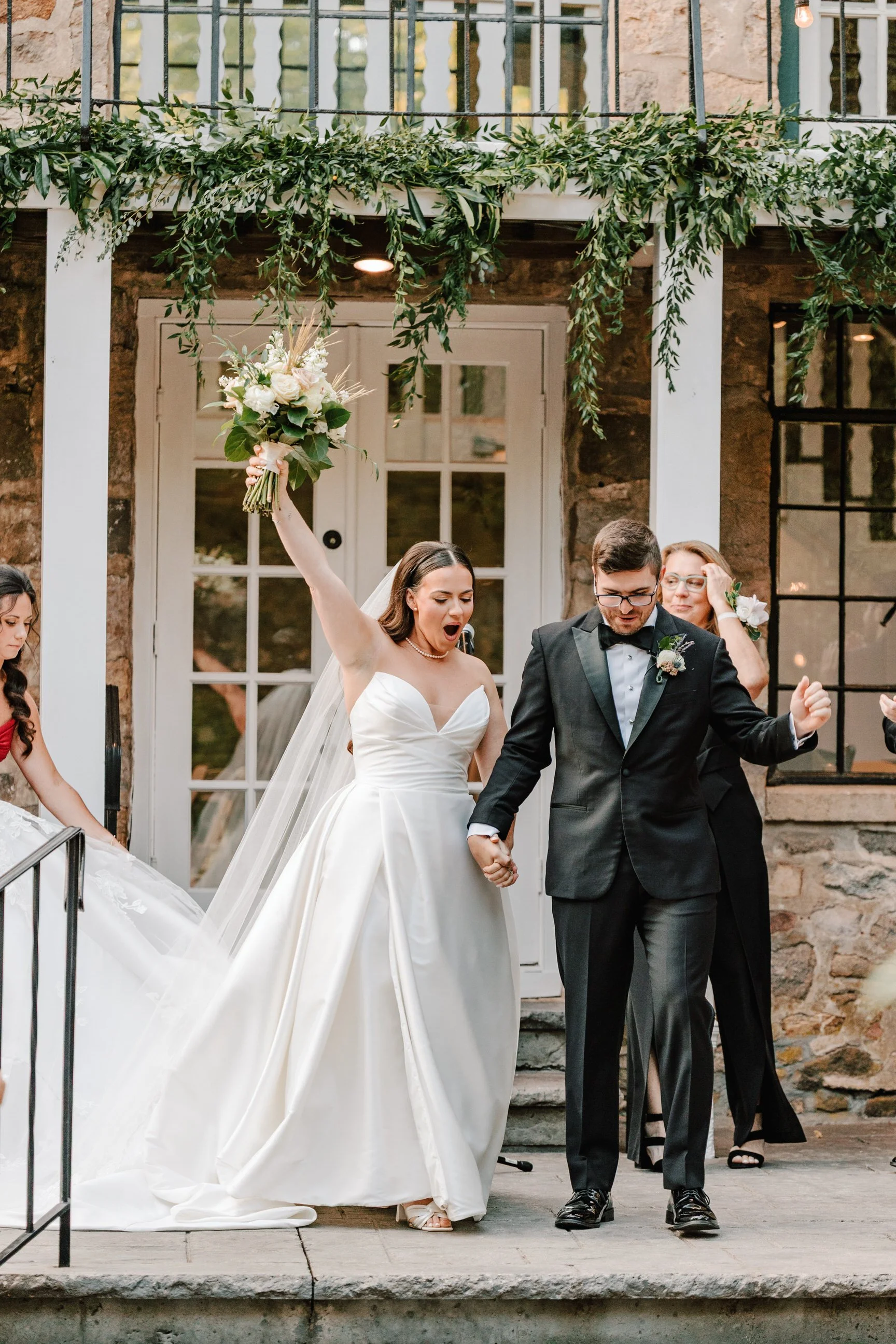 A bride in a white wedding dress celebrates with her arm raised, holding a bouquet, holding hands with a groom in a black tuxedo during their wedding ceremony outside a stone building.