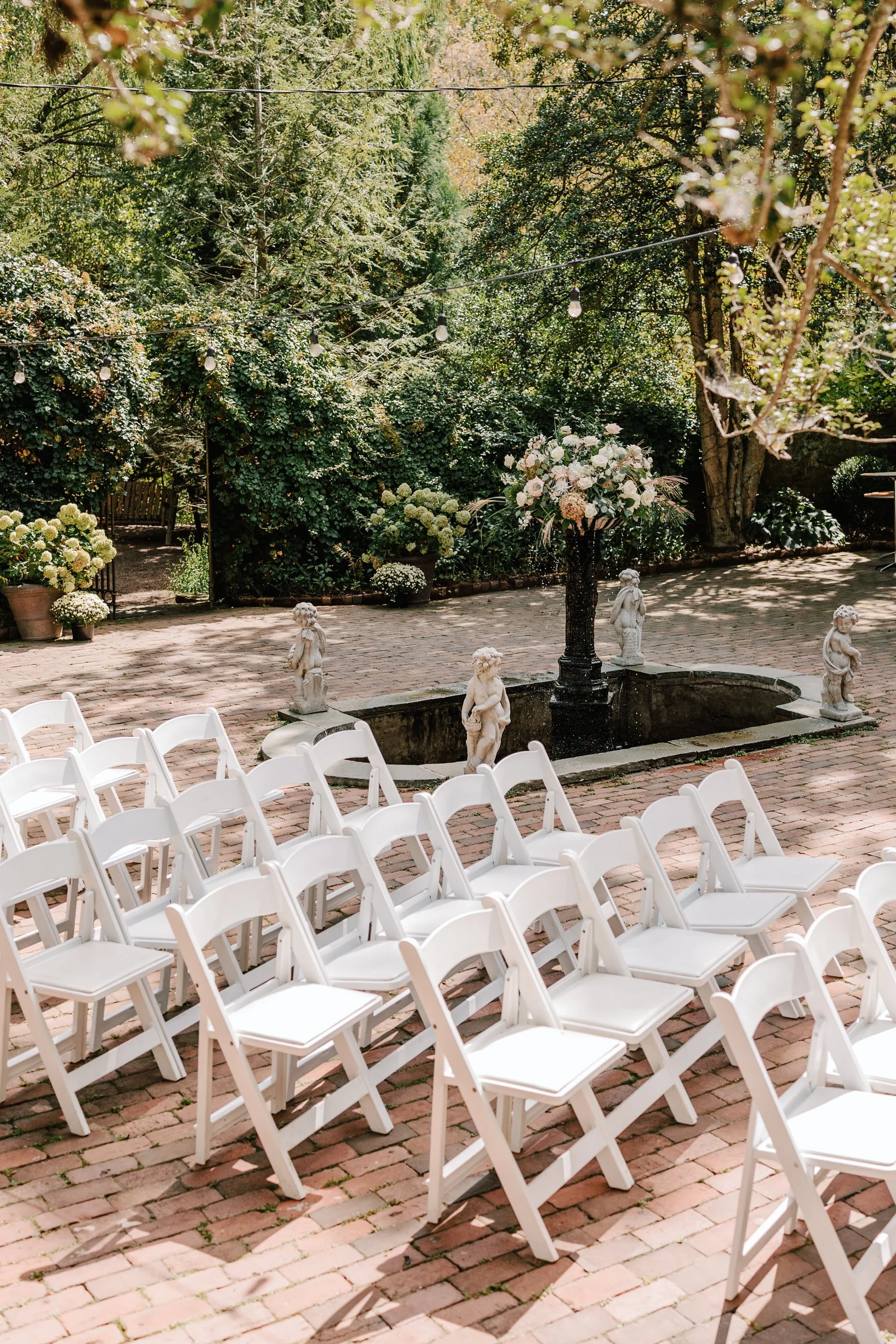 Outdoor wedding seating arrangement with white chairs facing a small fountain with statues and a large floral centerpiece, surrounded by lush greenery and trees.