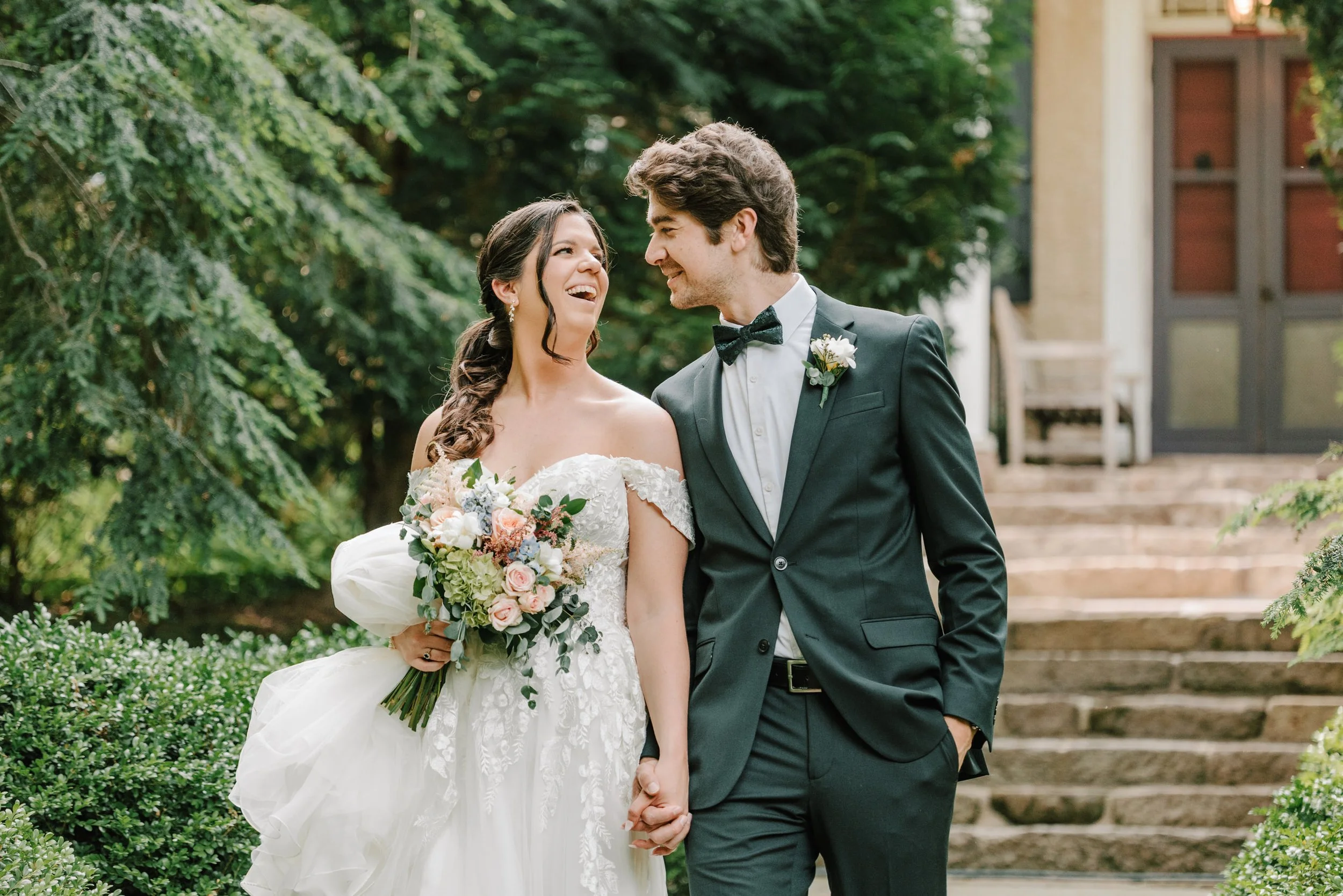A bride and groom holding hands and smiling at each other outdoors on a wedding day, with trees and steps in the background.