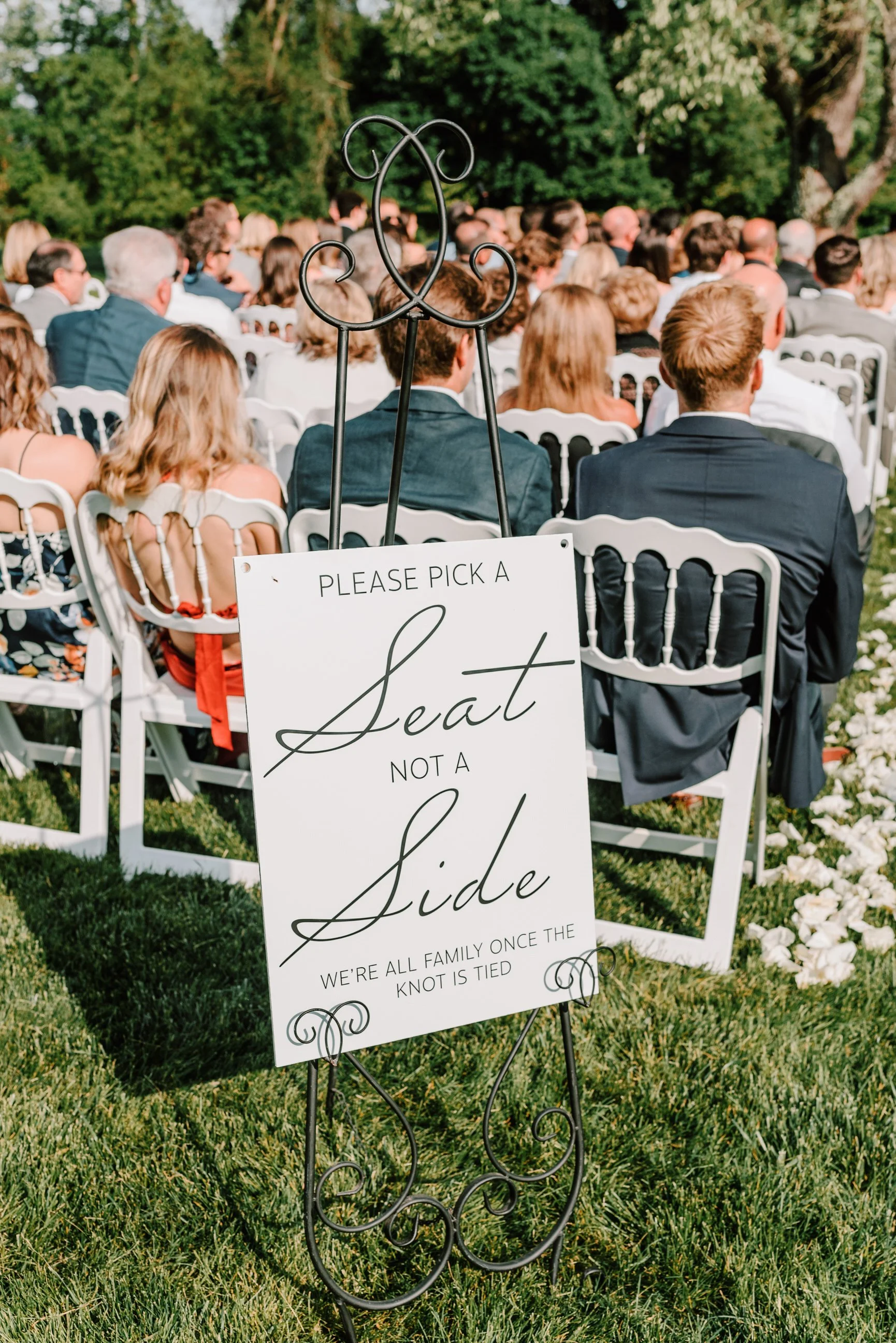Wedding ceremony with guests seated outdoors on white chairs; a sign in the foreground reads, 'Please pick a seat not a side. We're all family once the knot is tied.' Green trees and sunlight are visible in the background.