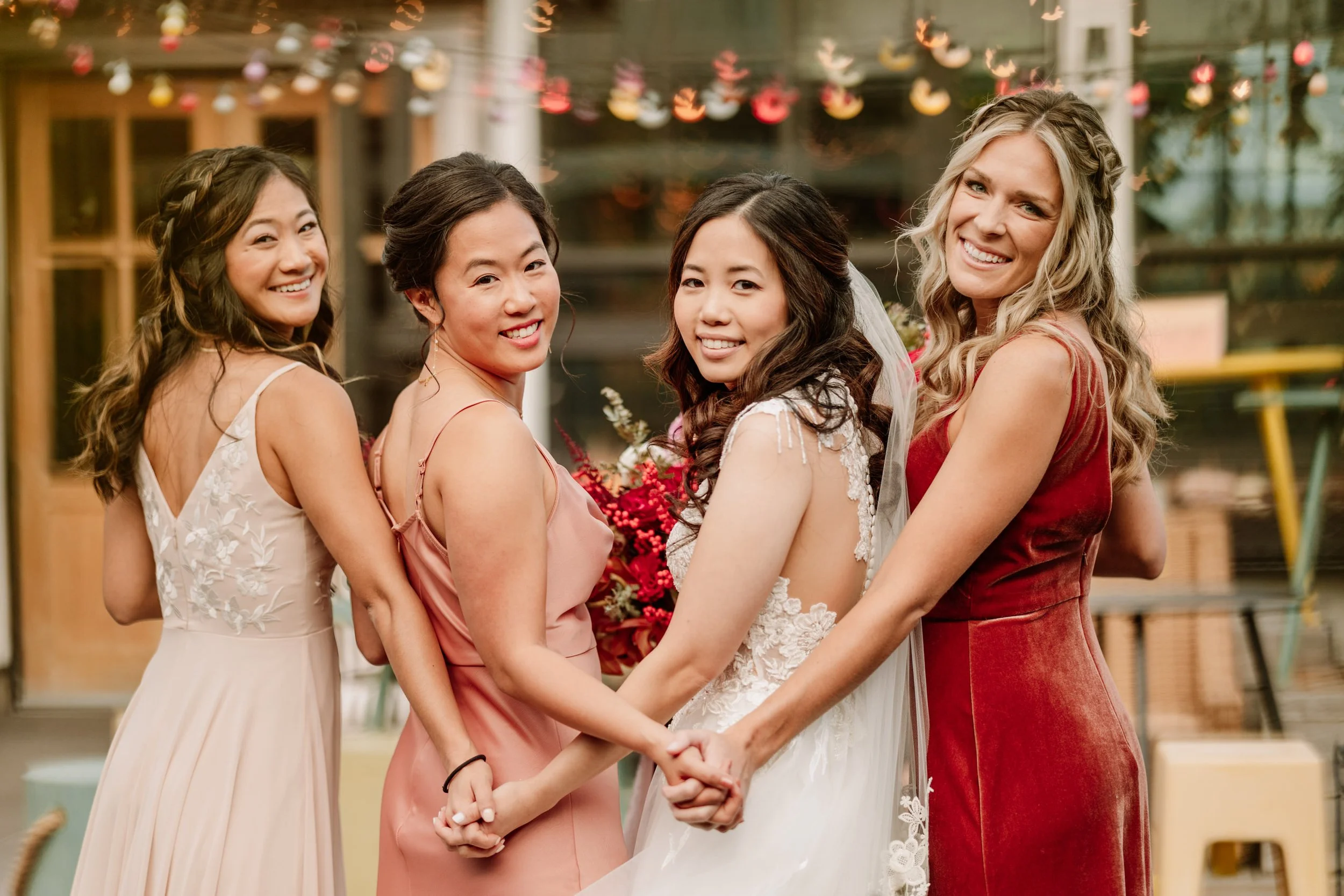 Four women at a wedding, including the bride wearing a white dress and veil, holding hands, smiling, with colorful lights and decorations in the background.