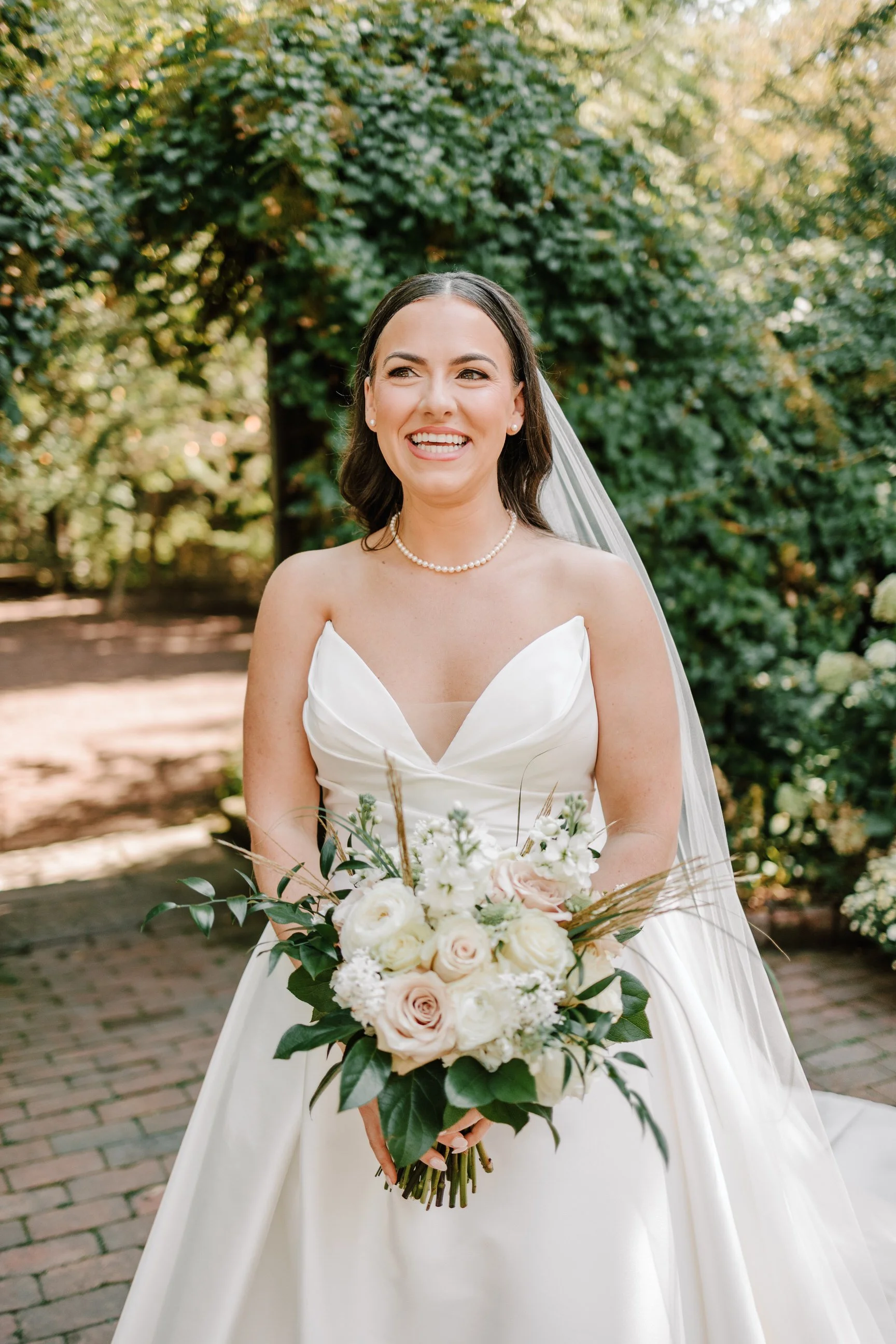A smiling bride in a white strapless wedding gown holding a bouquet of white and blush roses, greenery, and dried grasses outdoors with lush green trees in the background.