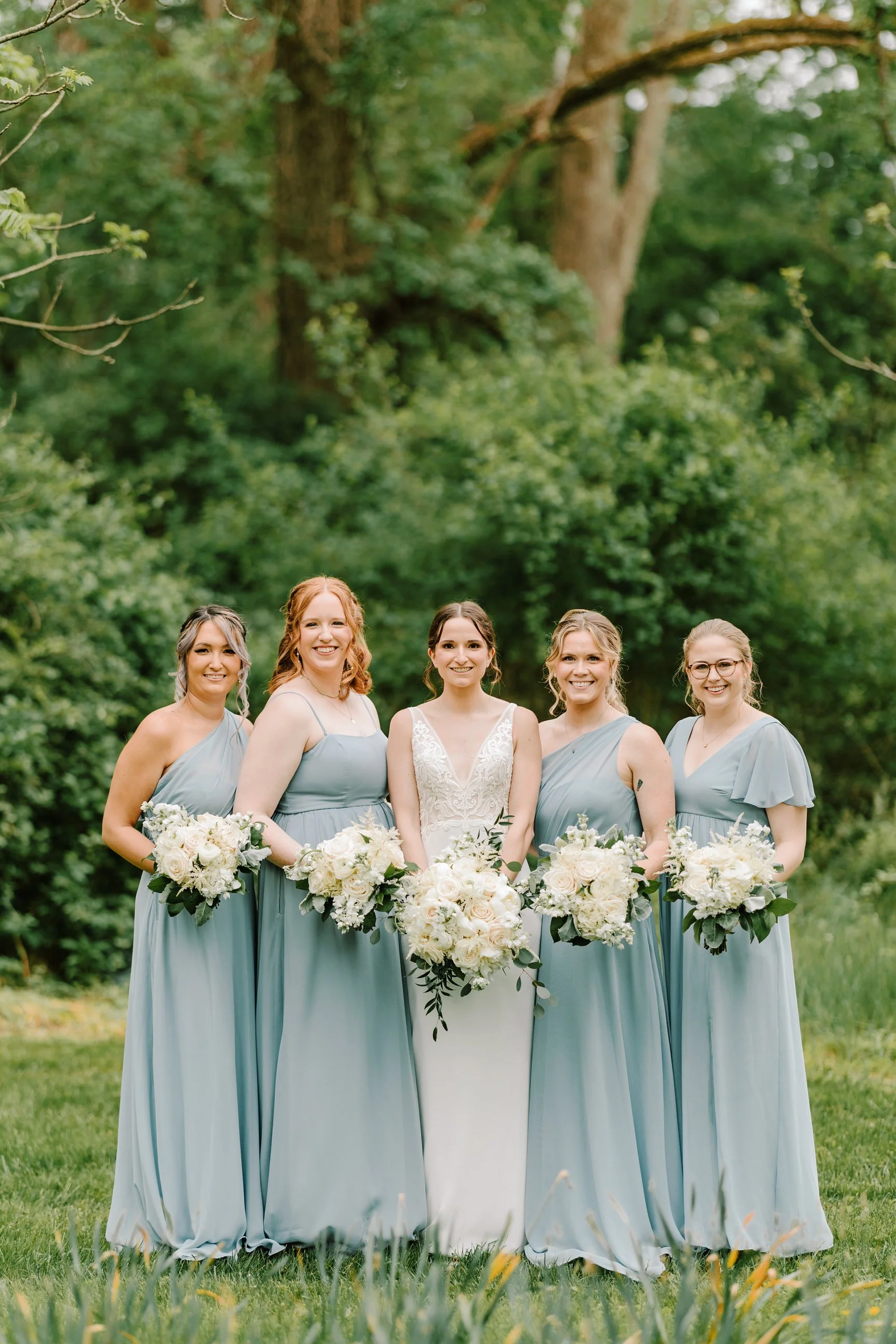 Group of five women, including a bride in a white dress and four bridesmaids in light blue dresses, standing outdoors in a lush green setting holding bouquets of white flowers.