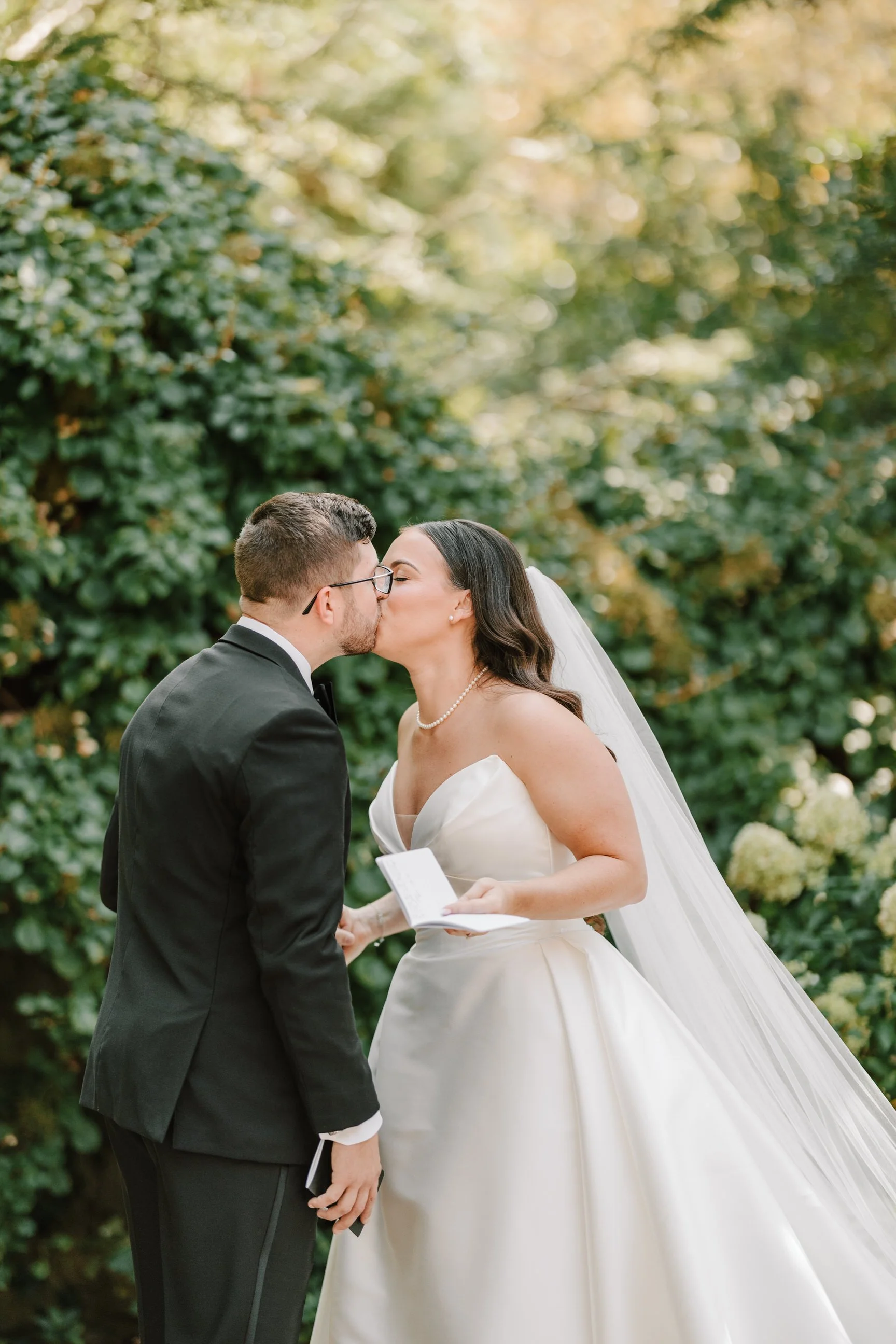 A bride and groom share a kiss during their wedding ceremony outdoors, with lush greenery in the background.