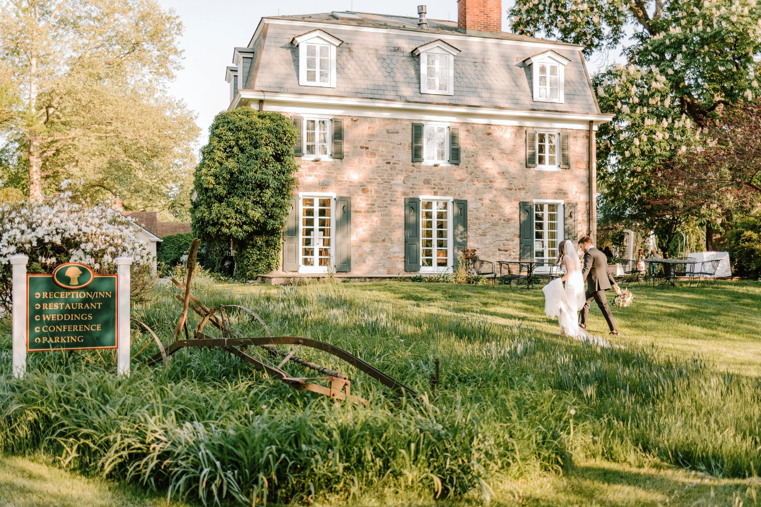 A bride and groom walking on a grassy lawn in front of a large stone house with multiple windows and green shutters. There is a sign to the left listing amenities, and trees and outdoor furniture are visible around the house, suggesting a wedding or 
