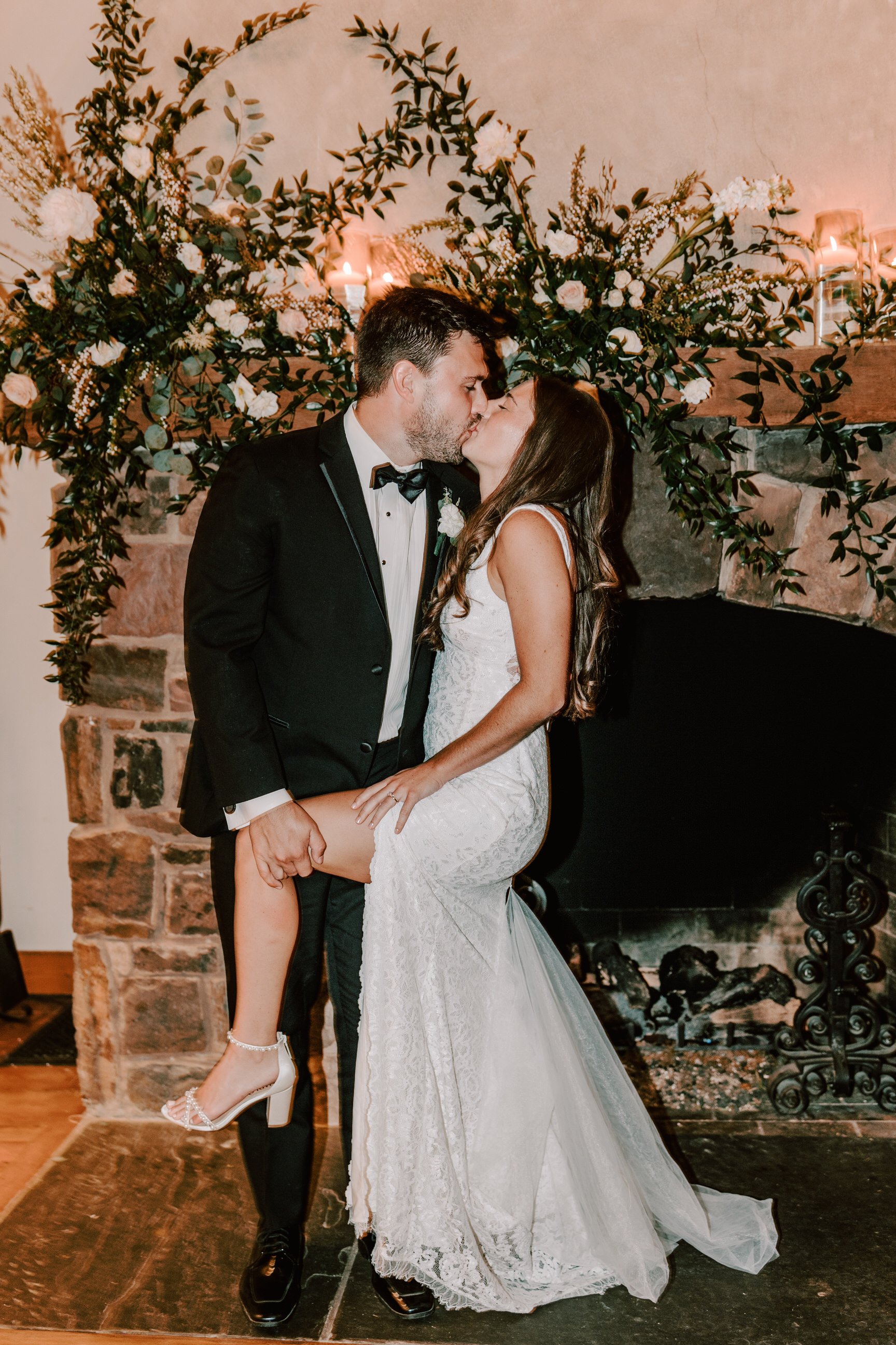 A bride and groom sharing a kiss during their wedding celebration. The groom is wearing a tuxedo and the bride is in a white lace wedding dress, with the bride lifting one leg while kissing. They are standing in front of a decorated stone fireplace w