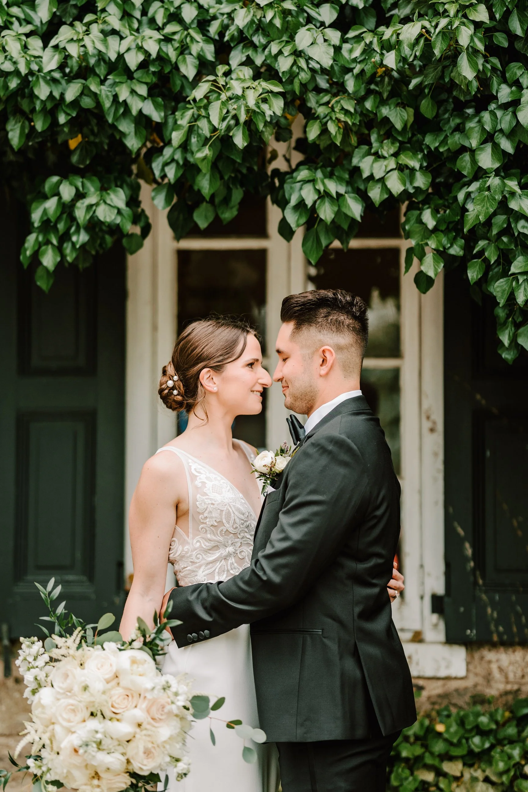A bride and groom standing close together, smiling, in front of a vintage wooden house with green shutters and lush green foliage.