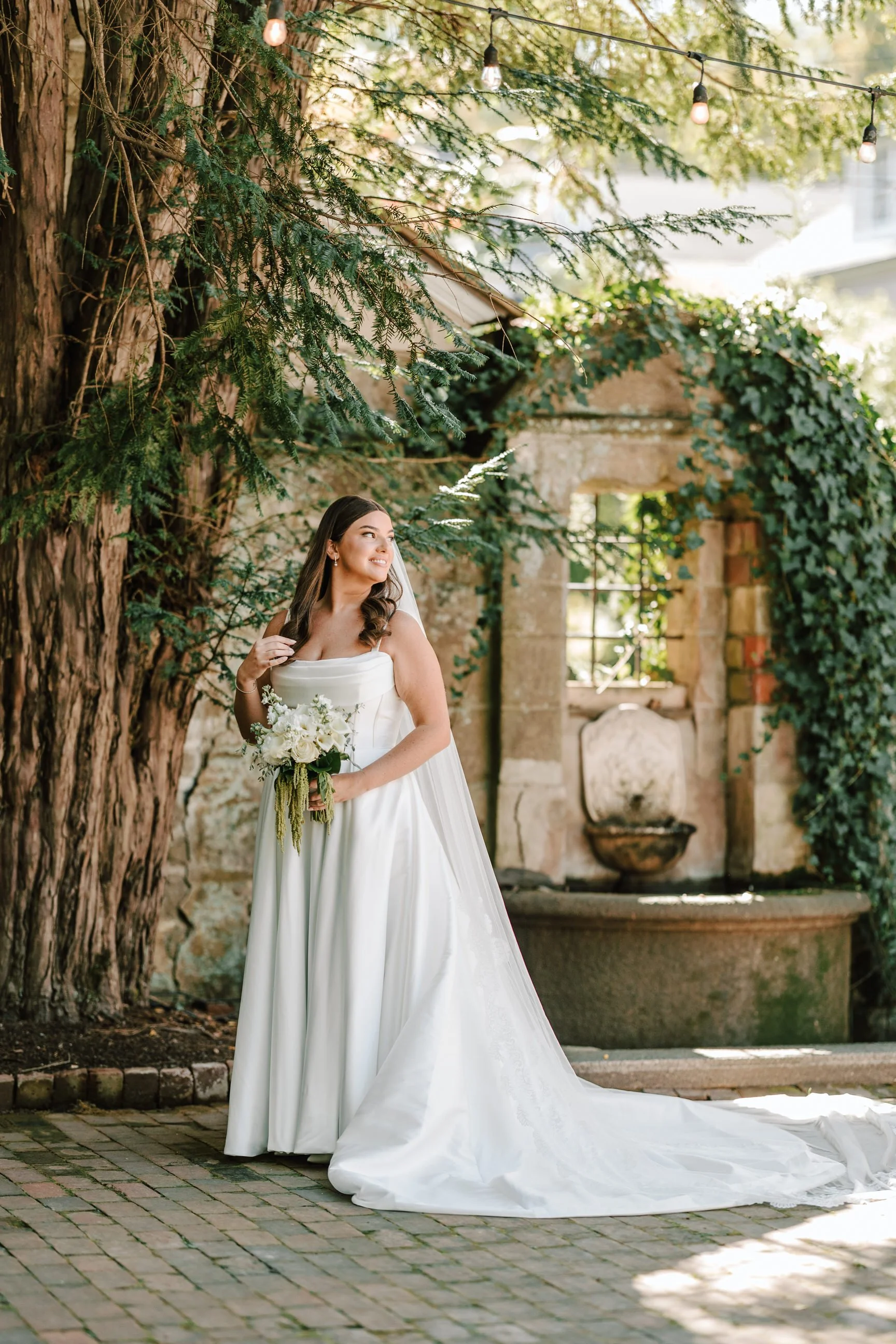 A bride standing outdoors near a large tree, holding a bouquet of white flowers, wearing a white wedding gown, with a stone fountain and ivy-covered stone wall in the background.