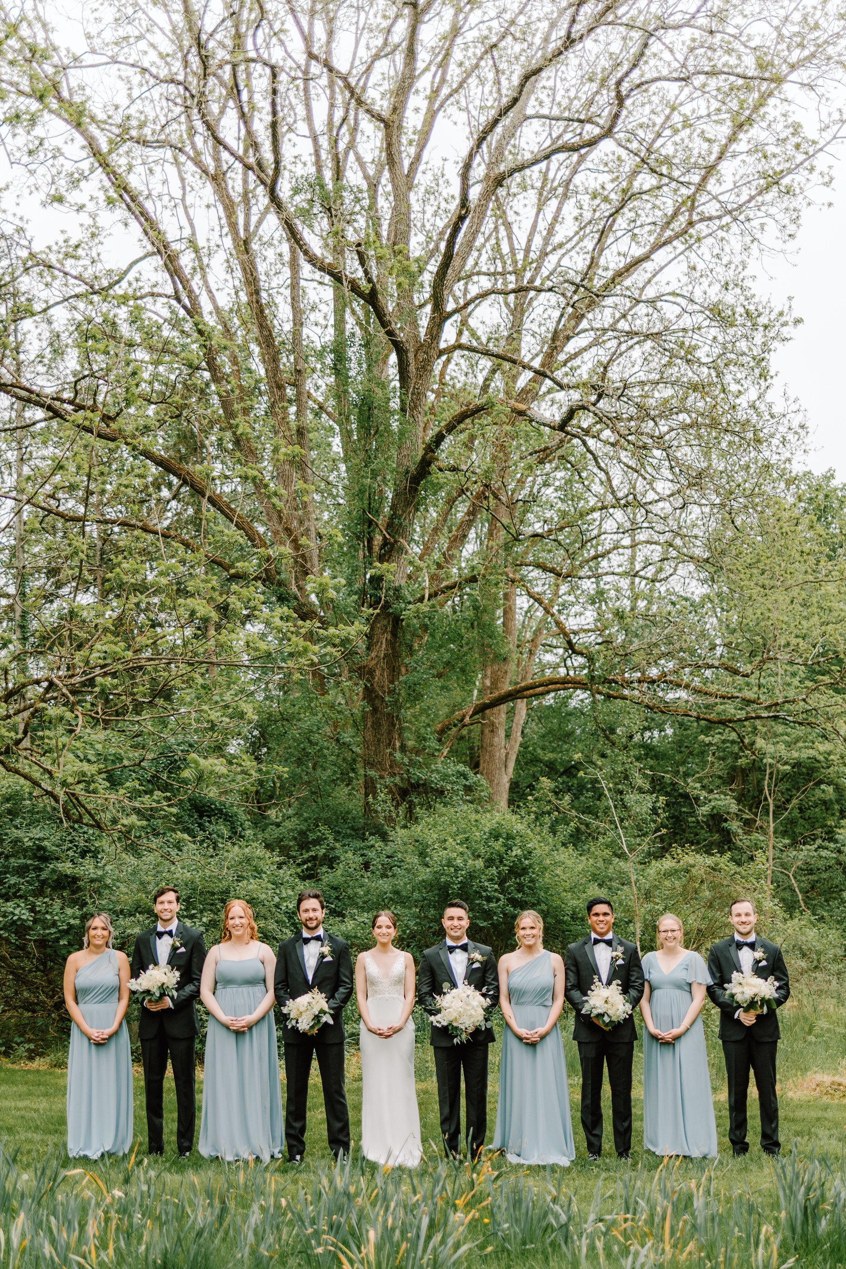 A wedding party of ten people standing outdoors on a grassy area in front of a large tree and green foliage. The group includes six women in light blue dresses and four men in black tuxedos with bowties, some of whom are holding white flower bouquets