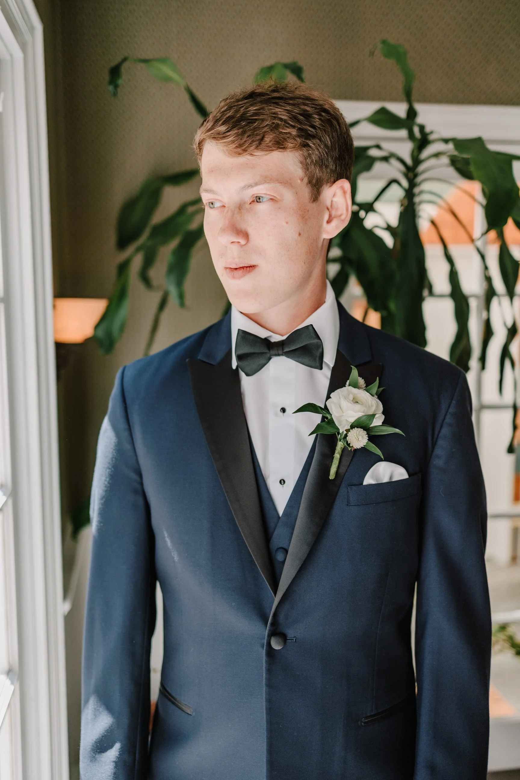 A young man in a tuxedo with a boutonniere stands by a window, looking outside, with a large green plant behind him.