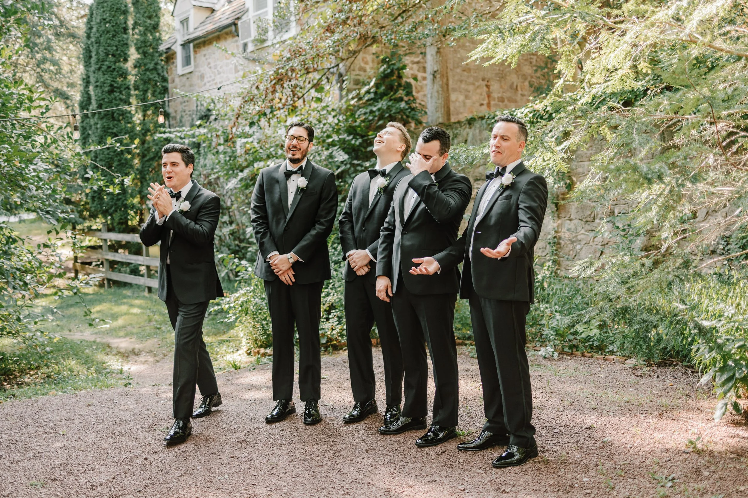 Six men dressed in black tuxedos with white shirts and black bow ties, standing outdoors on a gravel pathway, with a brick building and lush green trees in the background. They are engaged in a joyful moment, some laughing, some wiping their faces, w