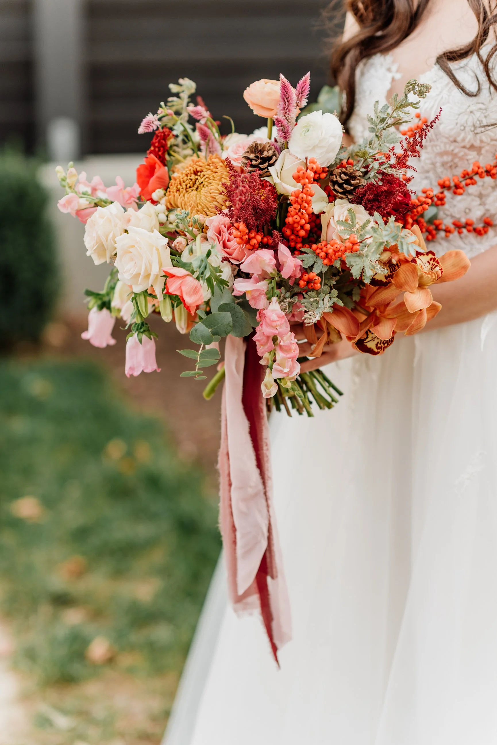 A person holding a colorful bouquet of various flowers including pink, red, orange, and white blooms, along with green foliage and pinecones, with a white dress and an outdoor background.