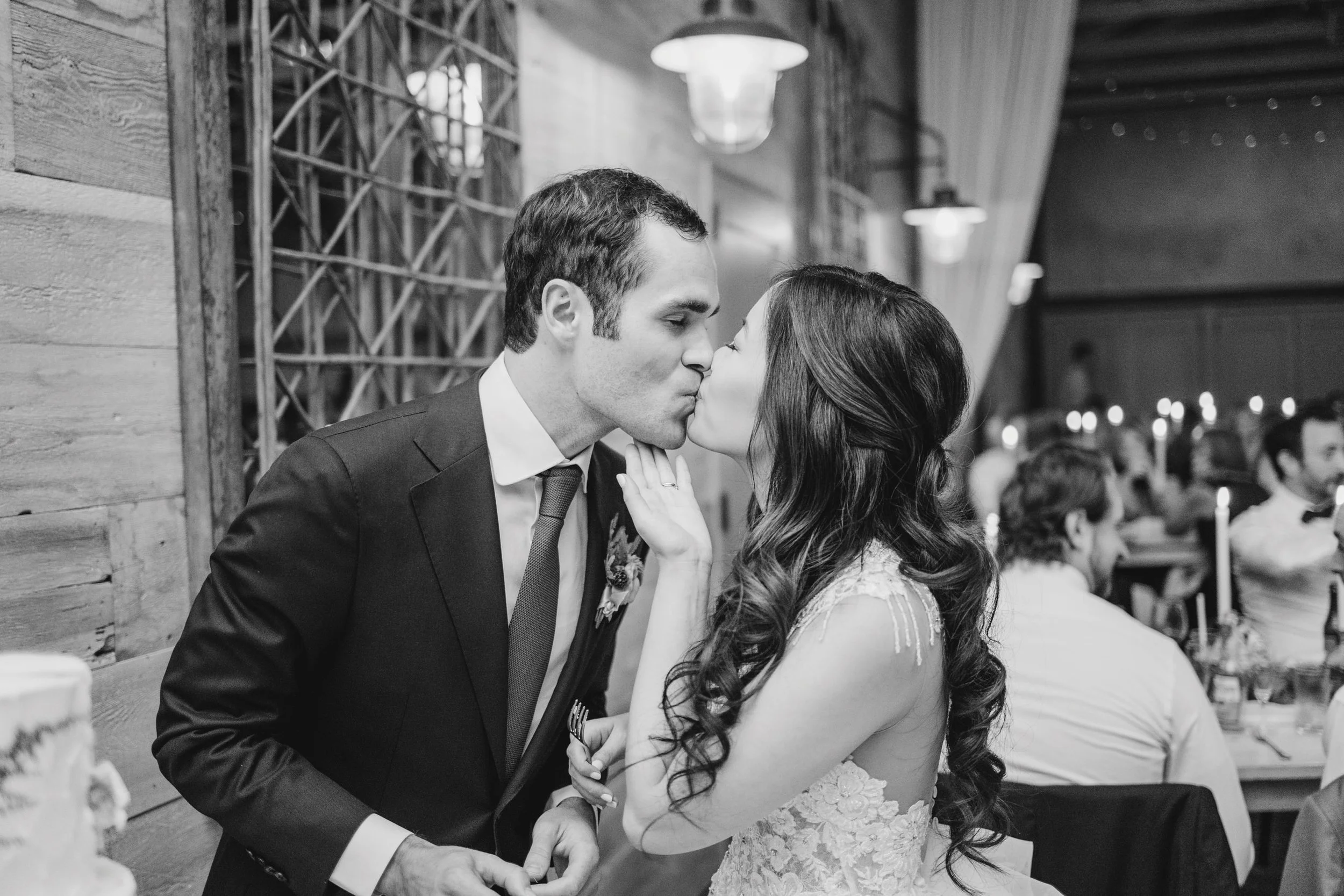 A black and white photo of a couple sharing a kiss at a wedding reception, with the bride in a lace dress and the groom in a suit and tie, in a decorated indoor venue.