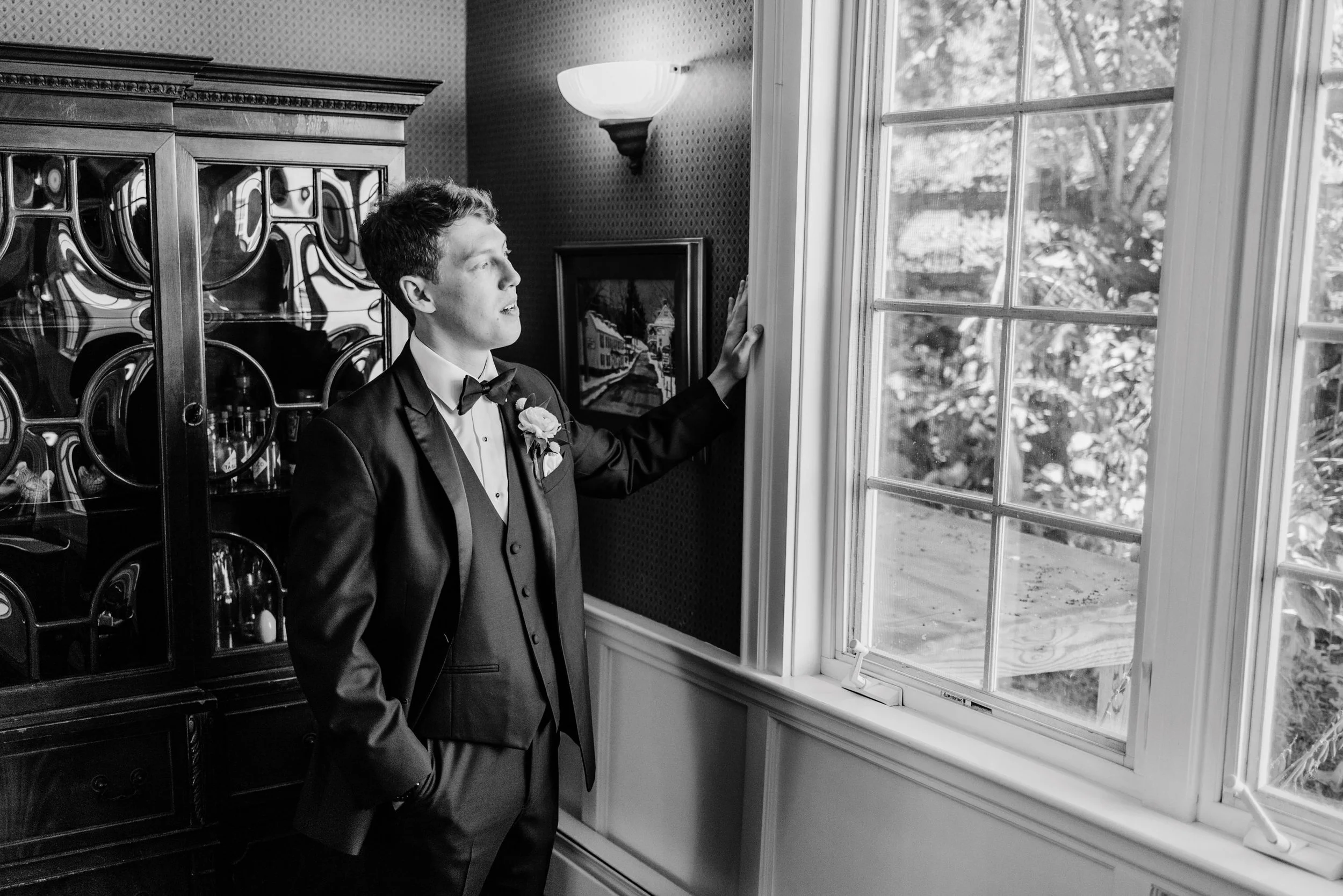 A young man in a tuxedo looking out a window, standing inside a room with a wooden cabinet and framed picture.