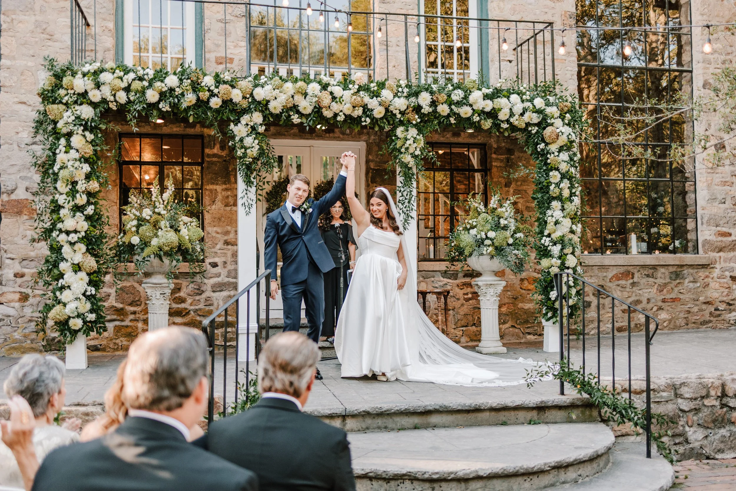 A newlywed couple celebrates their wedding victory as they stand on a decorated outdoor stone stage, holding hands and smiling after the ceremony. The bride wears a white wedding gown with a long train, and the groom wears a dark blue tuxedo. They ar